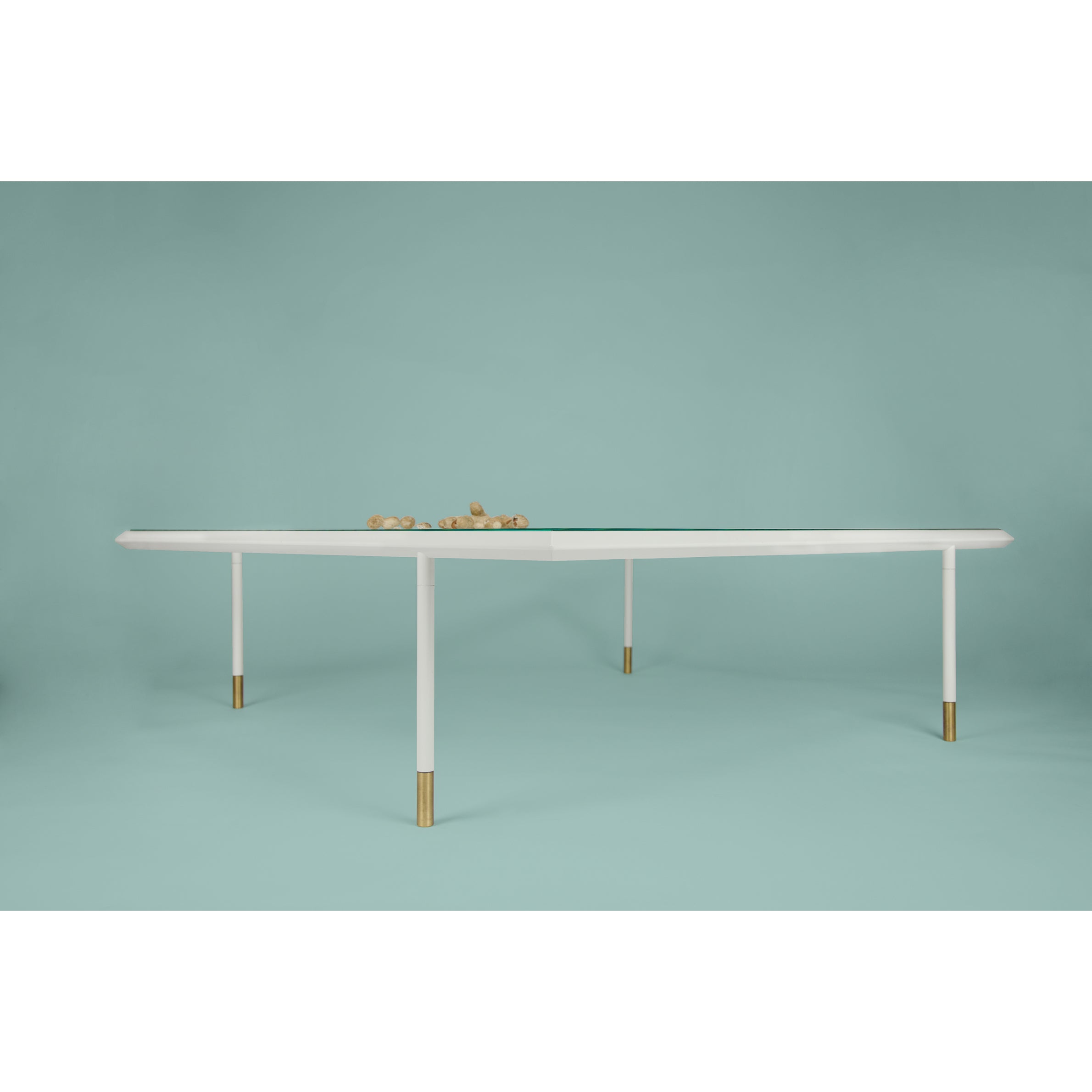 The Brutalesque Coffee Table by Coordination Berlin is a modern minimalist piece with a triangular glass top and white legs featuring gold accents. Set against a light teal background, it includes small decorative wooden elements on its surface.