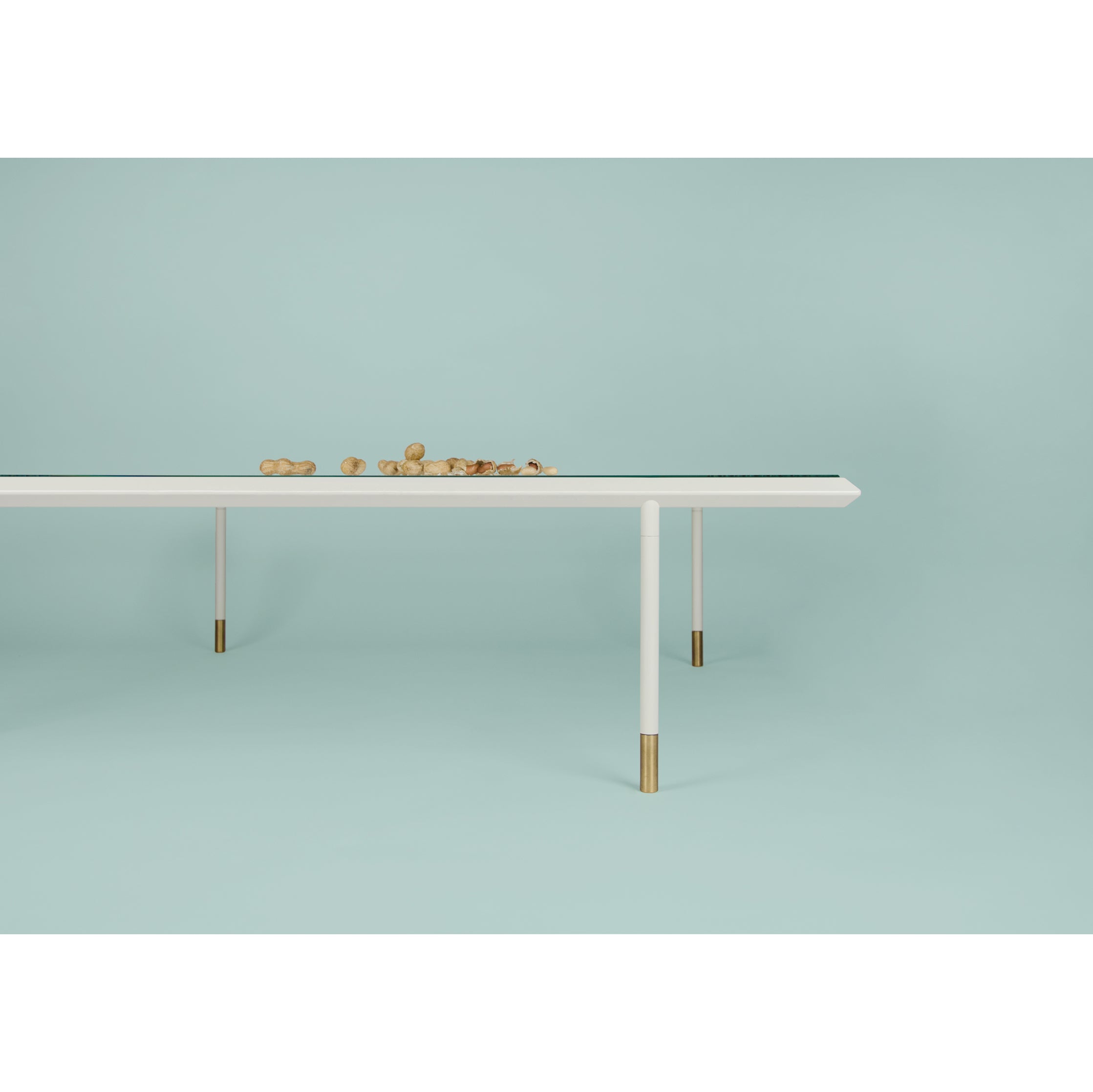 The Brutalesque Coffee Table by Coordination Berlin features a minimalist white surface with slim golden legs. Set against a light teal background, it exudes charm, complemented by wooden balls on top for a clean, modern aesthetic.