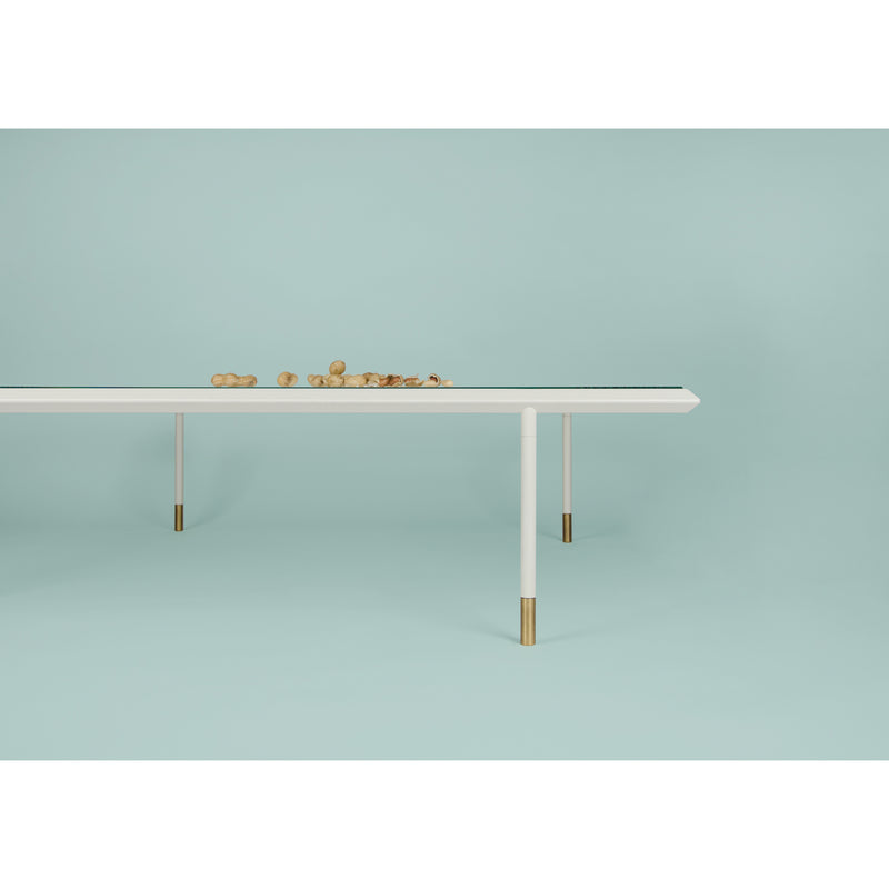 The Brutalesque Coffee Table by Coordination Berlin features a minimalist white surface with slim golden legs. Set against a light teal background, it exudes charm, complemented by wooden balls on top for a clean, modern aesthetic.