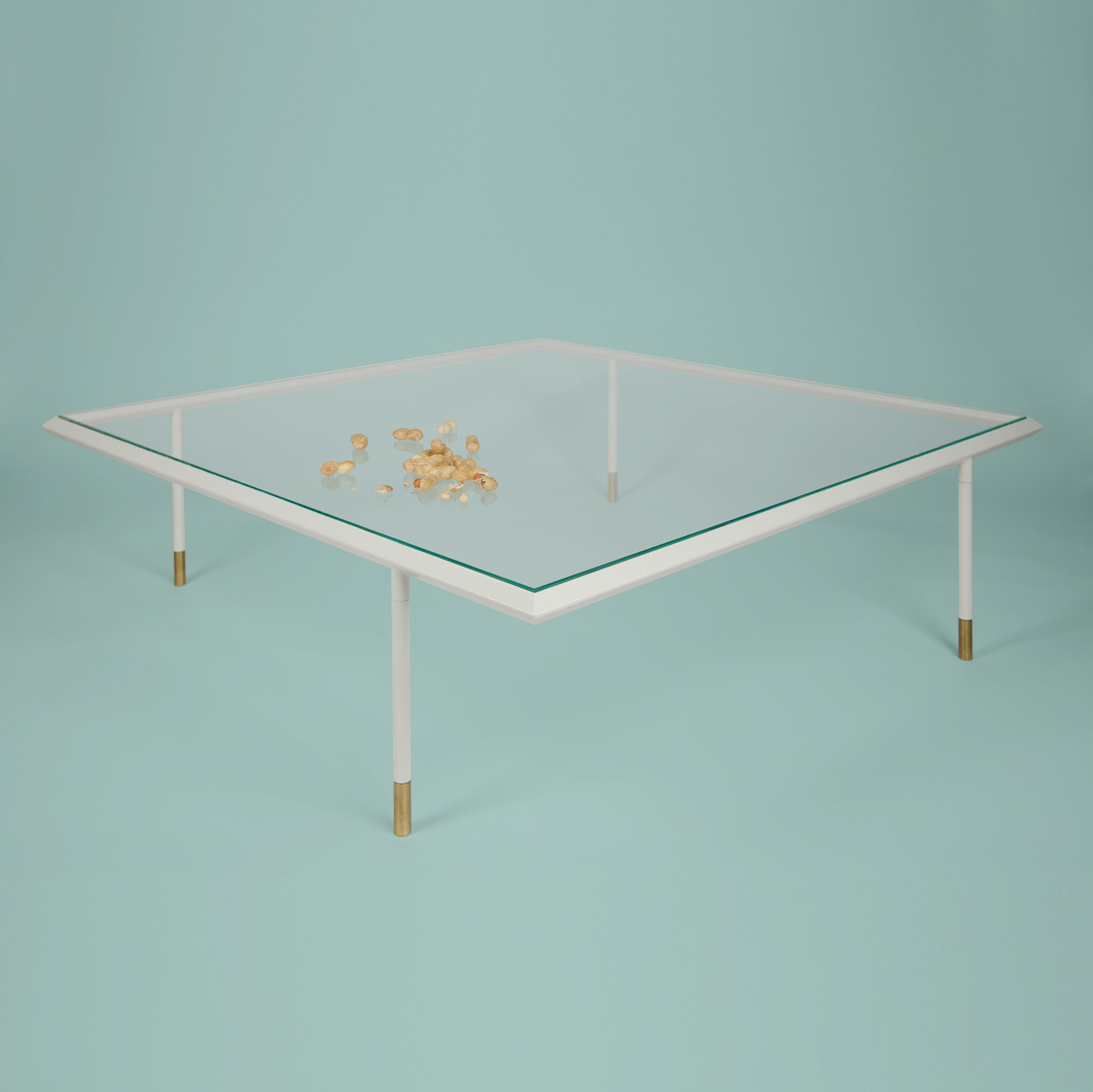 The Brutalesque Coffee Table by Coordination Berlin showcases a minimalist design with a glass top, white frame, and gold-tipped legs. Set against a light blue background, glinting gold coins are scattered across its surface to enhance its contemporary allure.