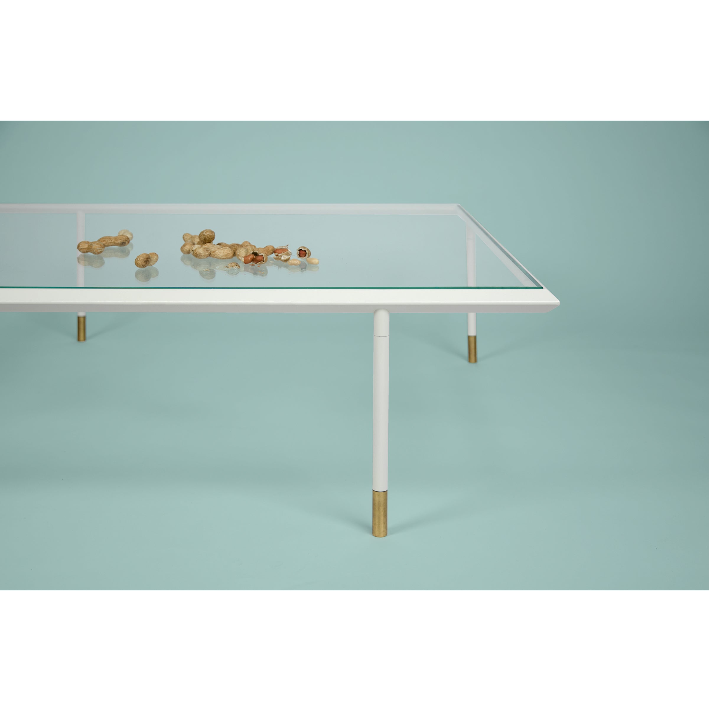 The Brutalesque Coffee Table by Coordination Berlin, featuring white legs and brass tips, stands against a blue backdrop. Its design subtly nods to Brutalist architecture as unshelled peanuts scattered on the tabletop introduce a casual element to this modern scene.