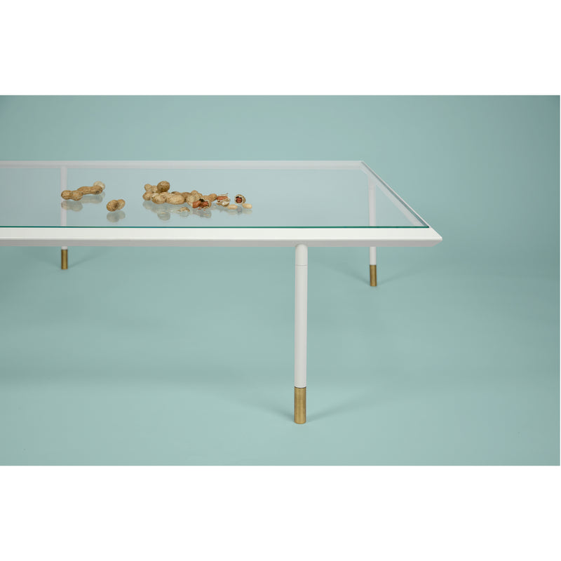 The Brutalesque Coffee Table by Coordination Berlin, featuring white legs and brass tips, stands against a blue backdrop. Its design subtly nods to Brutalist architecture as unshelled peanuts scattered on the tabletop introduce a casual element to this modern scene.