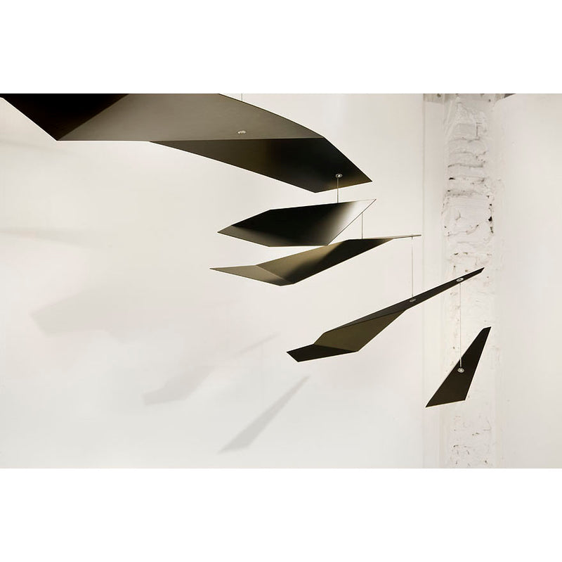 The Crowd mobile by Coordination Berlin features black geometric shapes suspended from the ceiling, varying in size and angle, casting shadows on a white wall. This creates a dynamic, minimalist composition reminiscent of contemporary architecture.