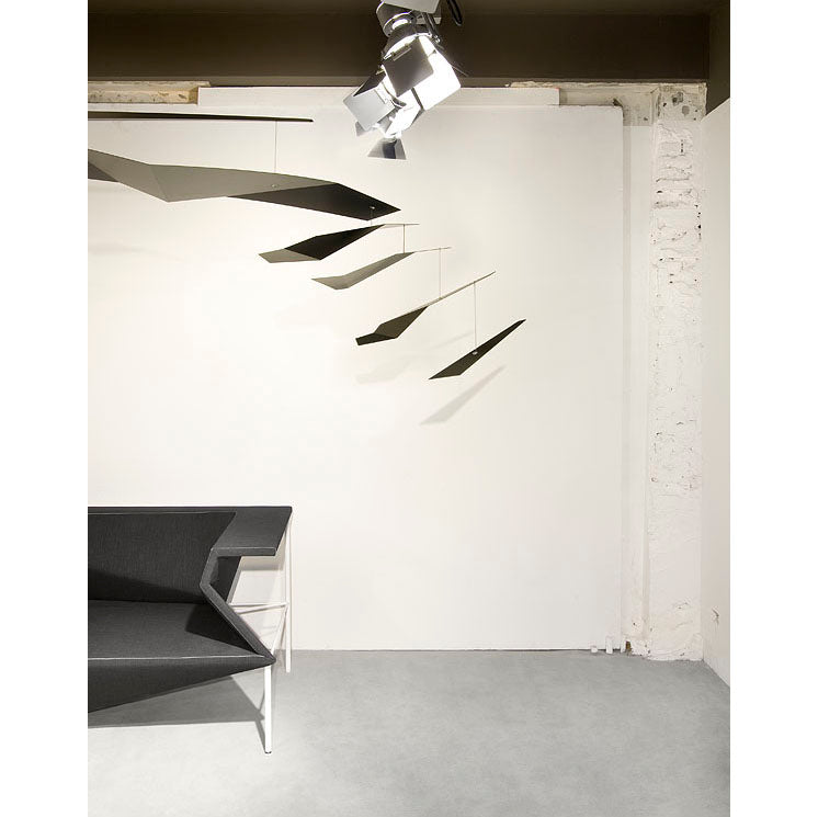 A minimalist room, echoing contemporary architecture, showcases a Coordination Berlins Crowd mobile in black by a white wall. Below it, an angular-designed black bench pairs with a small white table on the light gray floor, creating a harmonious ambiance.