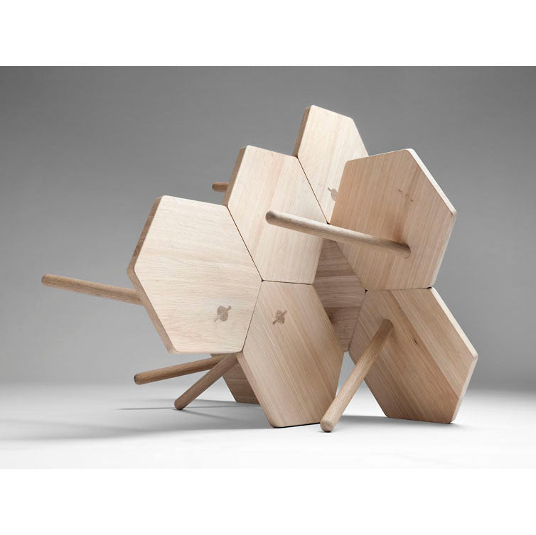 The Lean Tables by Coordination Berlin are crafted from local oak, featuring a hexagonal design with interlocking panels and cylindrical rods. Balanced at an angle, they showcase geometric patterns and a natural wood finish against a grey background, embodying contemporary design elements.