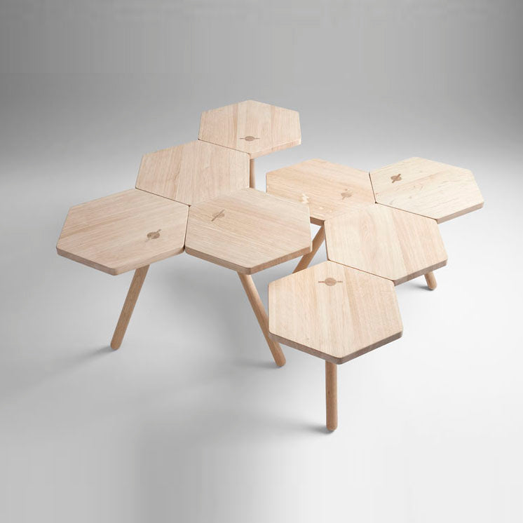 The Lean Tables by Coordination Berlin are interlocking hexagonal tables crafted from local oak. They feature a sleek, minimalist design with three legs, a light-colored finish, and are displayed in a honeycomb pattern on a light gray background.