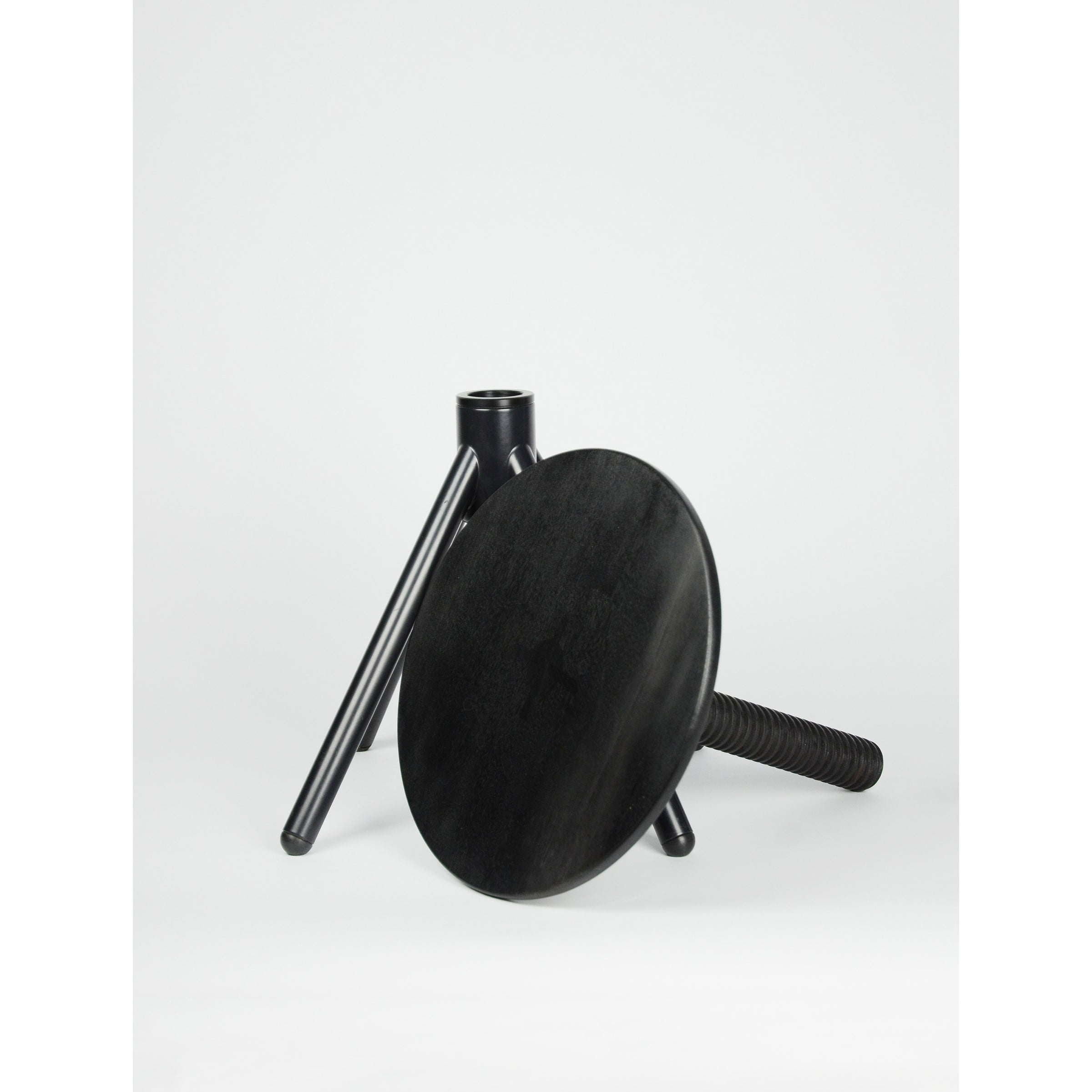 On a white background, a disassembled black wooden Thread Family - Stool by Coordination Berlin showcases its versatile design in walnut or oak, featuring three legs and a round seat. One leg is artfully leaned against the detached seat.