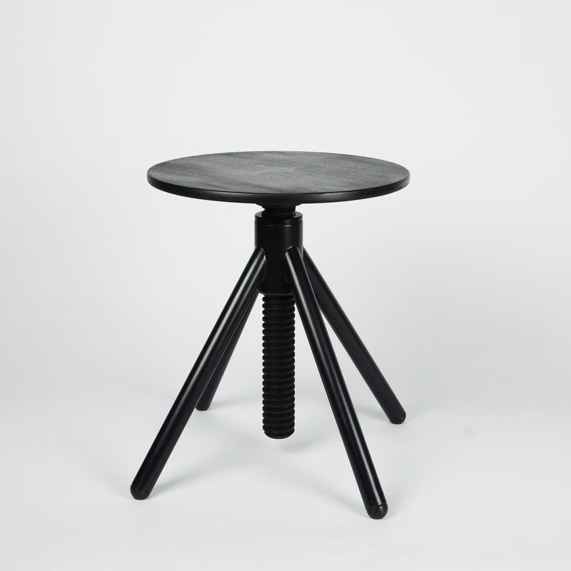 The Thread Family - Stool by Coordination Berlin is a minimalist black stool made from walnut wood, featuring a round seat, three slightly angled legs, and a central screw mechanism for adjusting height.