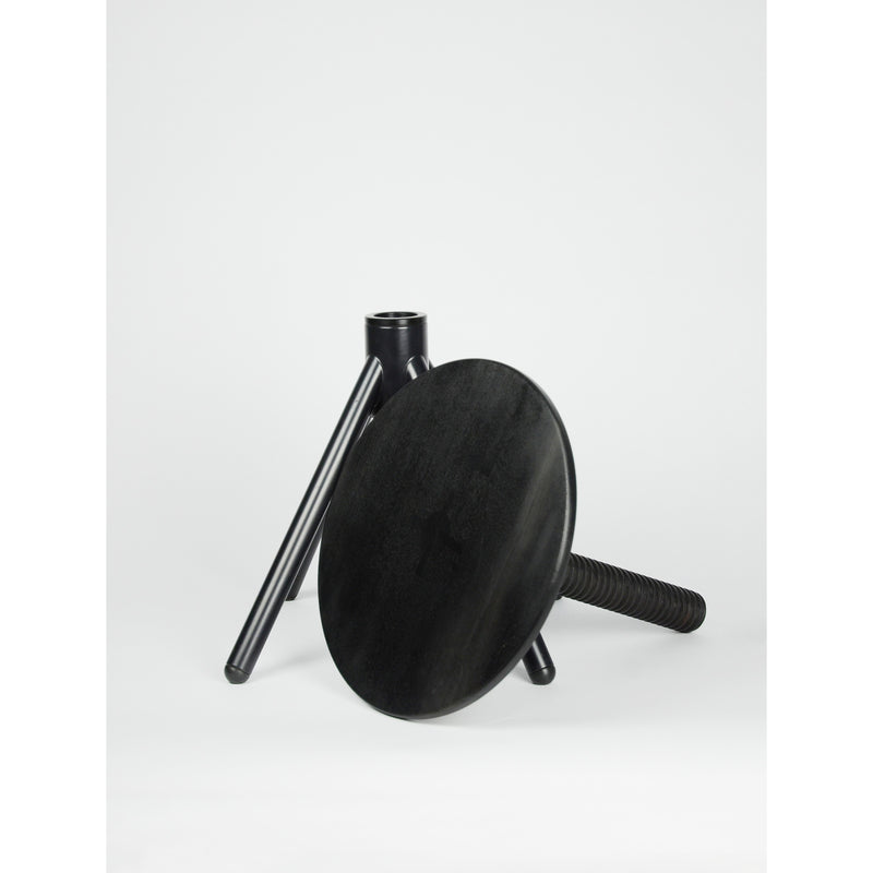 On a white background, a disassembled black wooden Thread Family - Stool by Coordination Berlin showcases its versatile design in walnut or oak, featuring three legs and a round seat. One leg is artfully leaned against the detached seat.