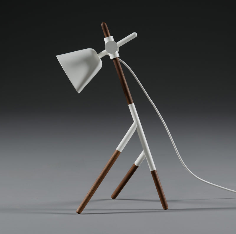 The Artisan INSERT Table Lamp features a minimalist design with a solid wood tripod base and a white cone-shaped shade against a gray background. It includes an eco-friendly white cord incorporating natural oils.