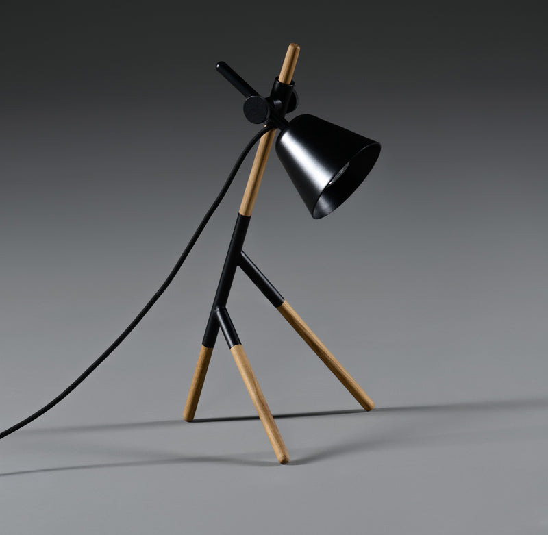 The Artisan INSERT Table Lamp features a modern tripod design with solid wood and black metal legs, complemented by a black conical shade and exposed cord. Set against a gray gradient background, it embodies an eco-friendly and minimalist aesthetic.