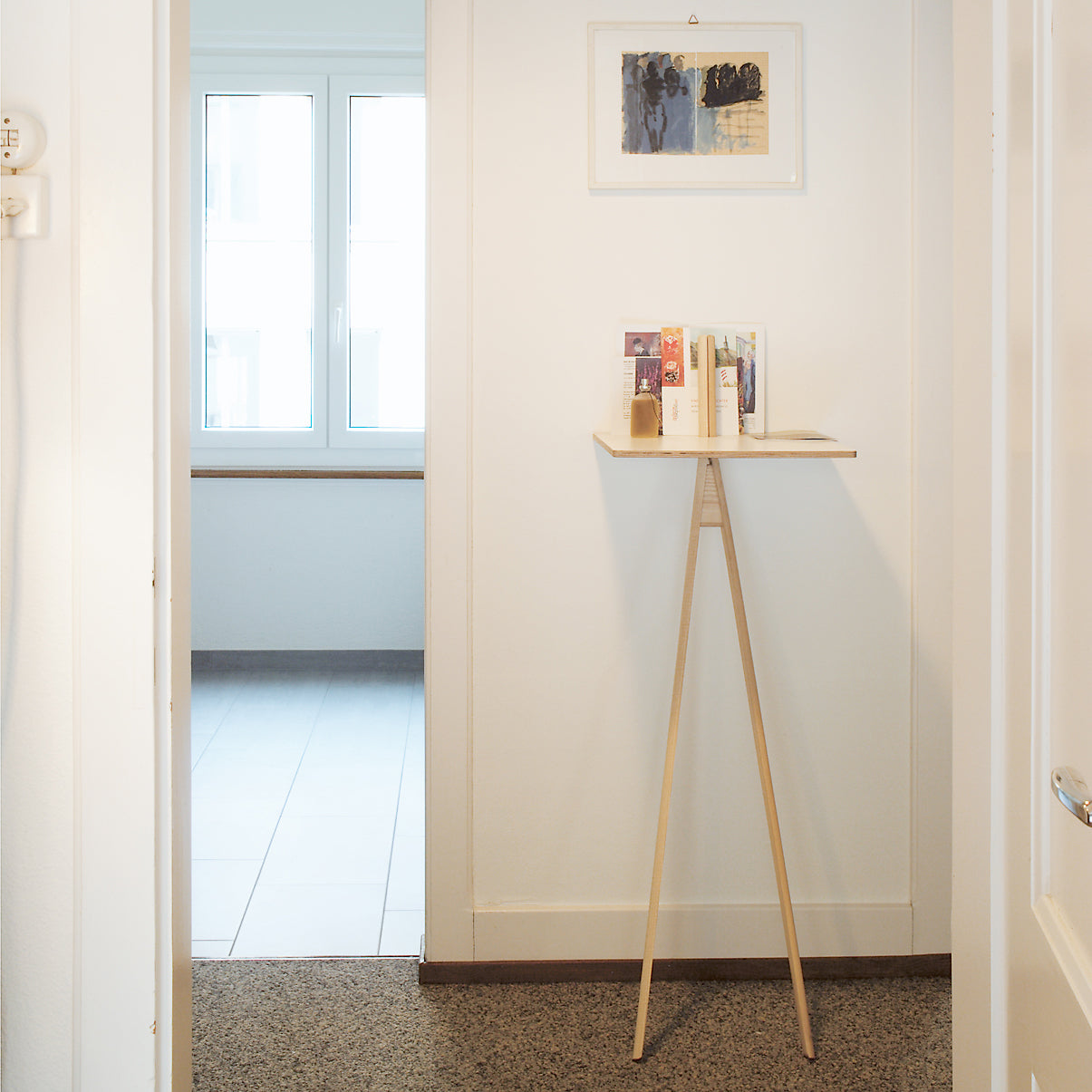 A minimalist hallway showcases a small wooden stand by Moormann, akin to Der Kleine Lehner, holding postcards, with framed art above. The efficient design leads into a bright room with a large window, transitioning the floor from carpet to tile.