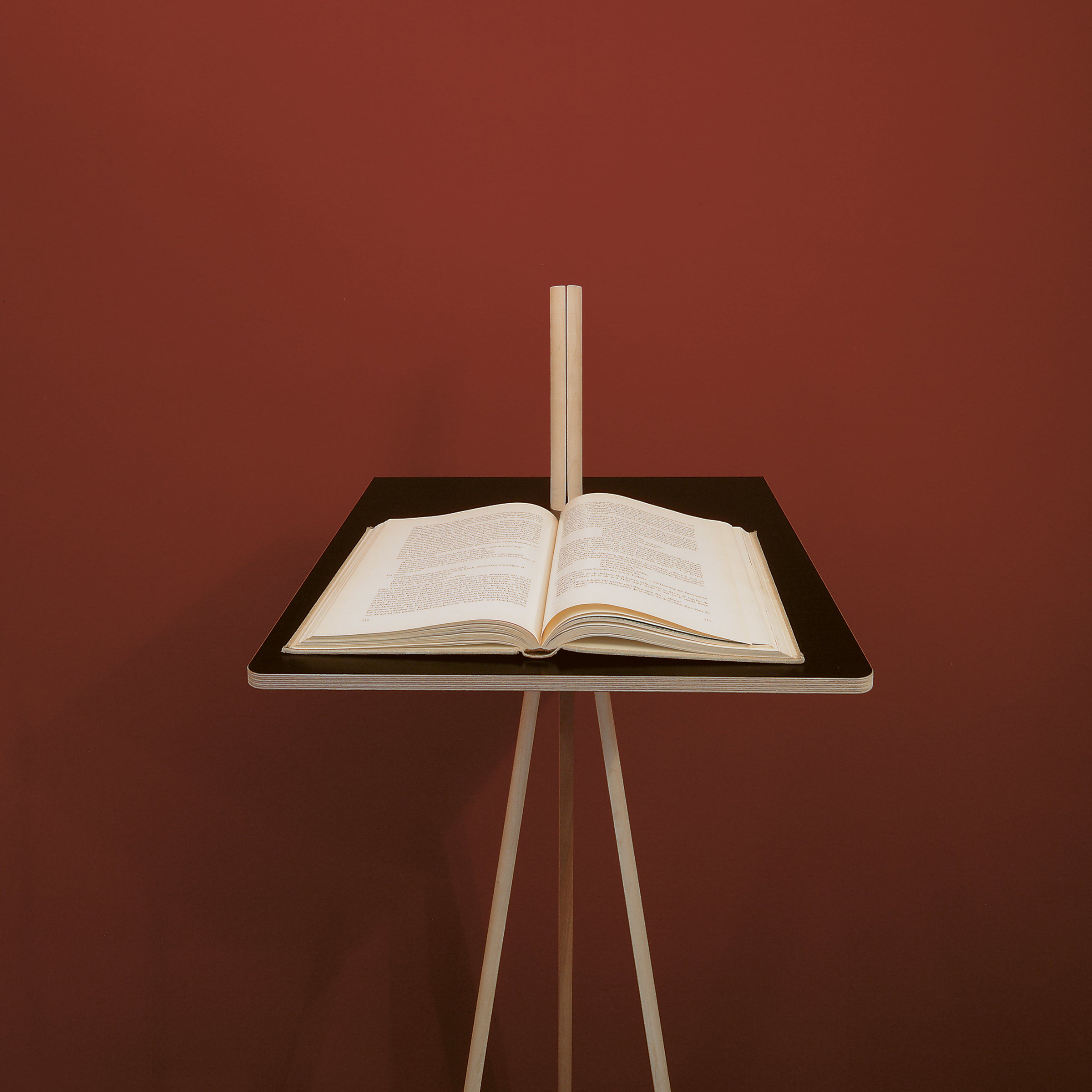 An open copy of Moormanns Der Kleine Lehner sits on a minimalist writing stand with light wooden legs, set against a maroon backdrop. The book is spread midway, displaying printed text on a stand with vertical back support for space-saving.