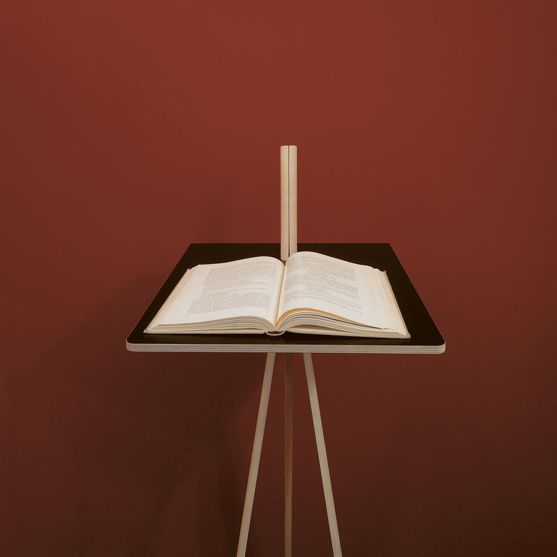 An open copy of Moormanns Der Kleine Lehner sits on a minimalist writing stand with light wooden legs, set against a maroon backdrop. The book is spread midway, displaying printed text on a stand with vertical back support for space-saving.