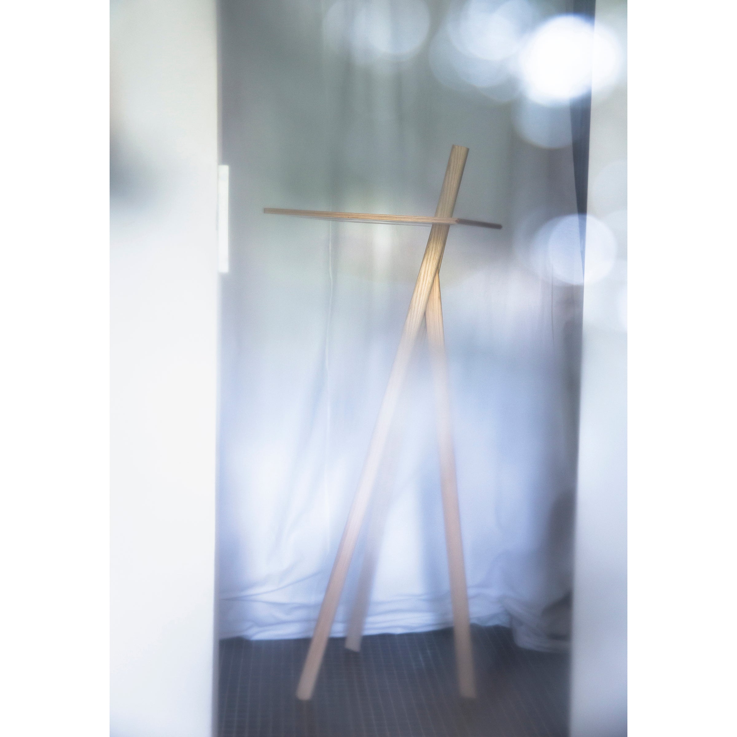 A minimalist wooden tripod easel from Moormann, called Der Kleine Lehner, stands against a softly lit gray fabric backdrop. The dreamy image with blurred circular light reflections in the upper right corner highlights its space-saving design and complements the serene ambiance beautifully.