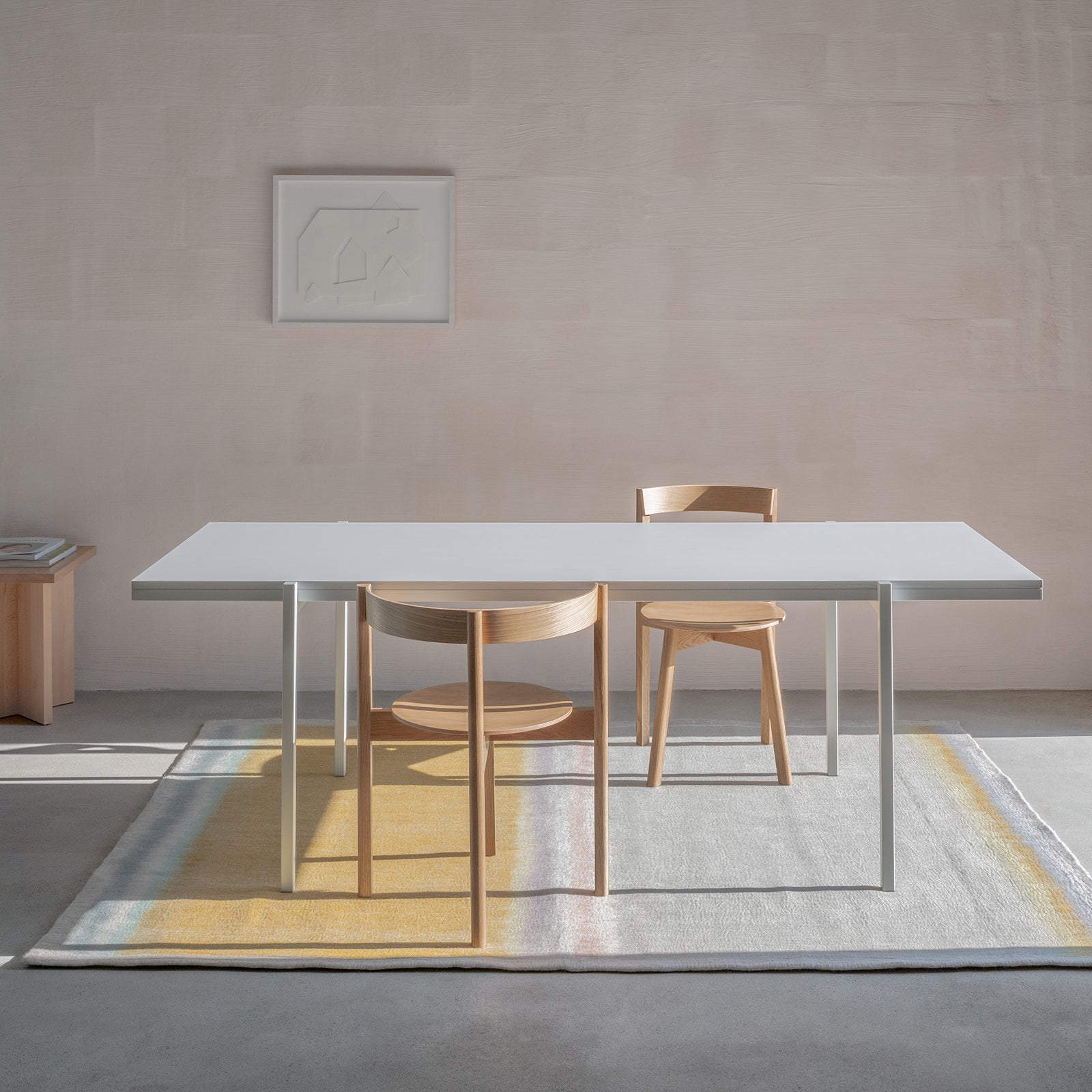 A minimalist room highlights a modern white table paired with two Loehr L5 JAZZ Armchairs of solid wood, set on a muted-textured rug. A framed abstract art piece adorns the light wall, with a wooden side table partially visible.
