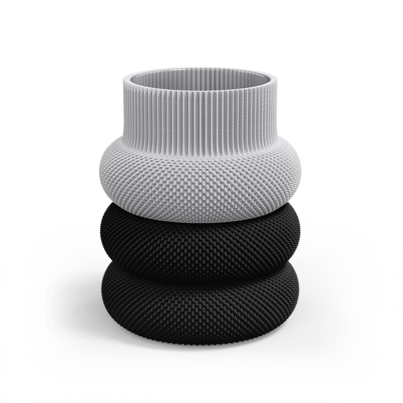 The Dorn 110g Vase by Sheyn is a modern, eco-friendly creation made via innovative 3D printing. It features a white, ribbed top and two sleek black sections below, with a rounded stacked design that contrasts in color and texture while resting on a reflective surface.
