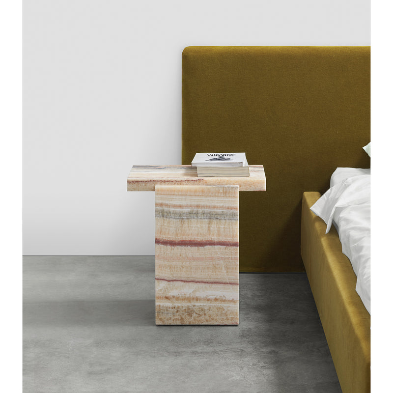 A modern bedroom features E15s DT02 TORE ONYX stone side table with stripes beside a mustard-yellow bed, evoking subtle Art Deco vibes. A stack of books on the table complements the minimalist design, characterized by David Thulstrups signature white and gray palette.