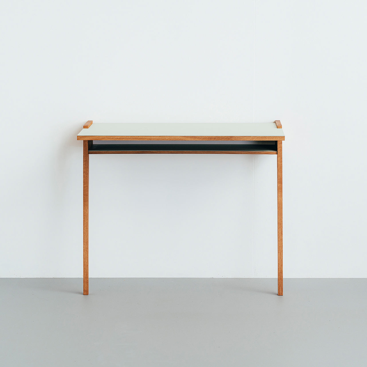 The Bachmann Desk by Das kleine b showcases a minimalist design with a light-colored solid oak top, slim legs against a white wall, and includes a discreet shelf beneath for functionality.