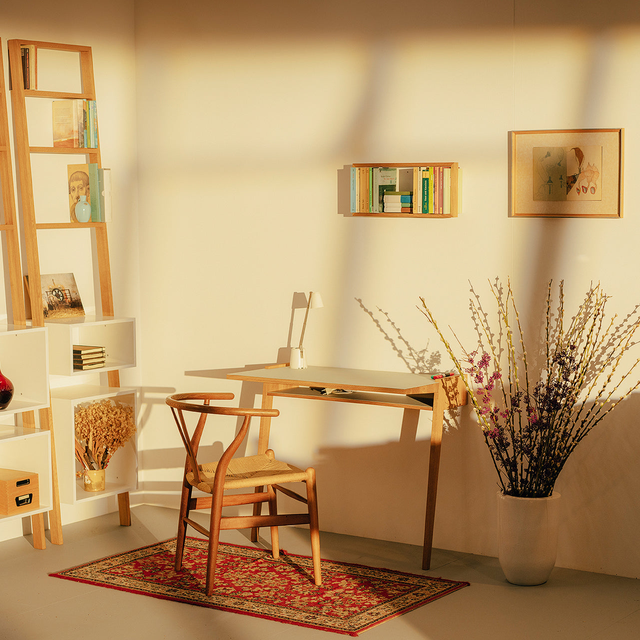 A sunlit minimalistic room showcases a Bachmann Desk by Das kleine b and a matching chair on a red patterned rug. Books and décor fill the shelves, while a wall hanging and large vase with branches and flowers create soft shadows on the white walls.