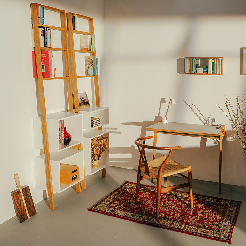 A minimalist home office features the Bachmann Desk by Das kleine b and a wooden chair on a decorative rug. Sunlight casts shadows on the white walls. A tall wooden shelf holds books and decor, with a small wall-mounted shelf and a plant branch nearby.