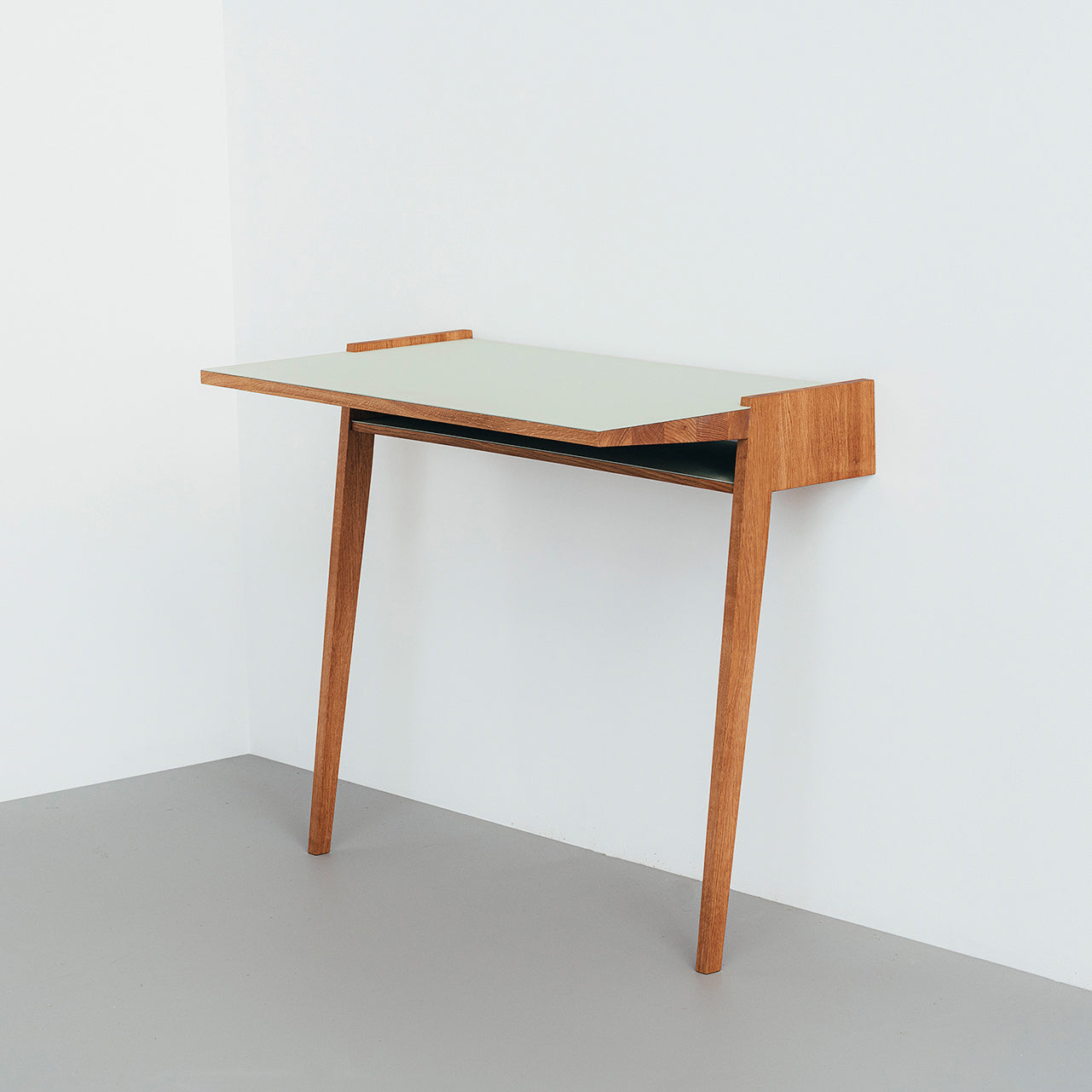 The Bachmann Desk by Das kleine b showcases a minimalist design with two front legs and a rear panel affixed to the wall. Its light-colored top complements the light gray floor and pale walls, fitting seamlessly into the corner.