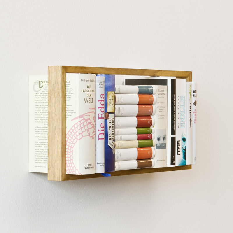 The Das kleine b 8.7 wall-mounted bookshelf features a wooden frame showcasing German books in a spiral around two bars. Some appear to float, adding intrigue with their varied colors and sizes, with titles and covers partly visible.