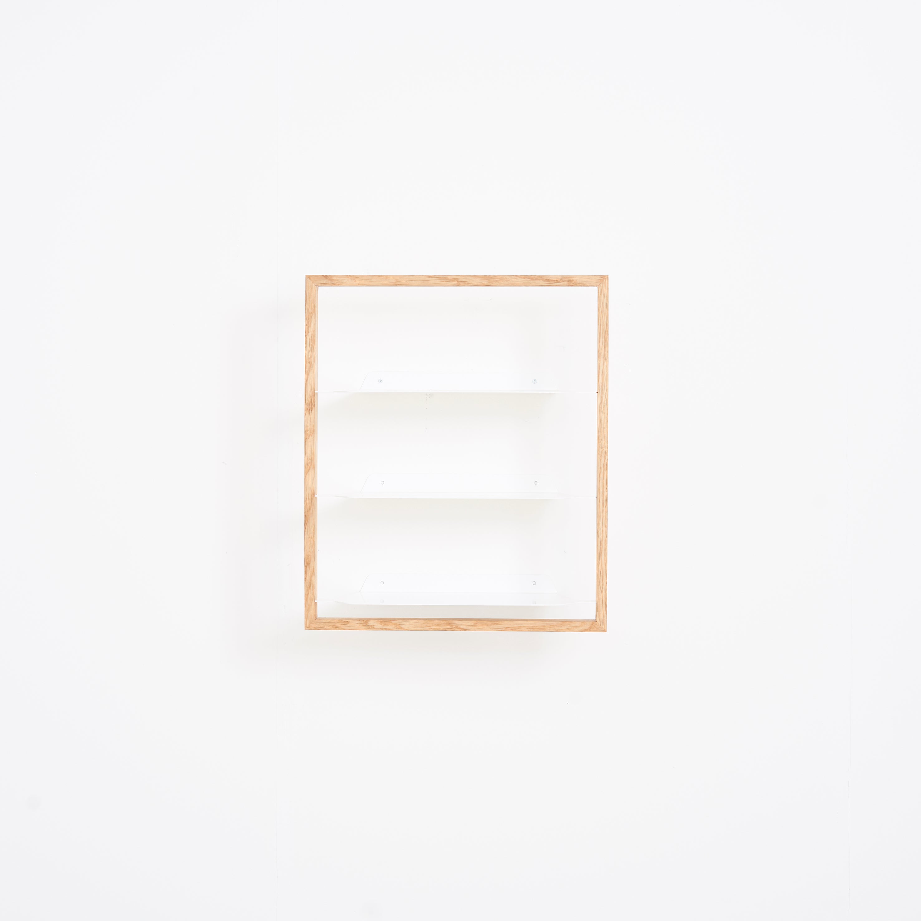The Basti by Das kleine b is a minimalist shelf with an oak frame mounted on a white wall, featuring three empty white compartments for a simple and clean design.