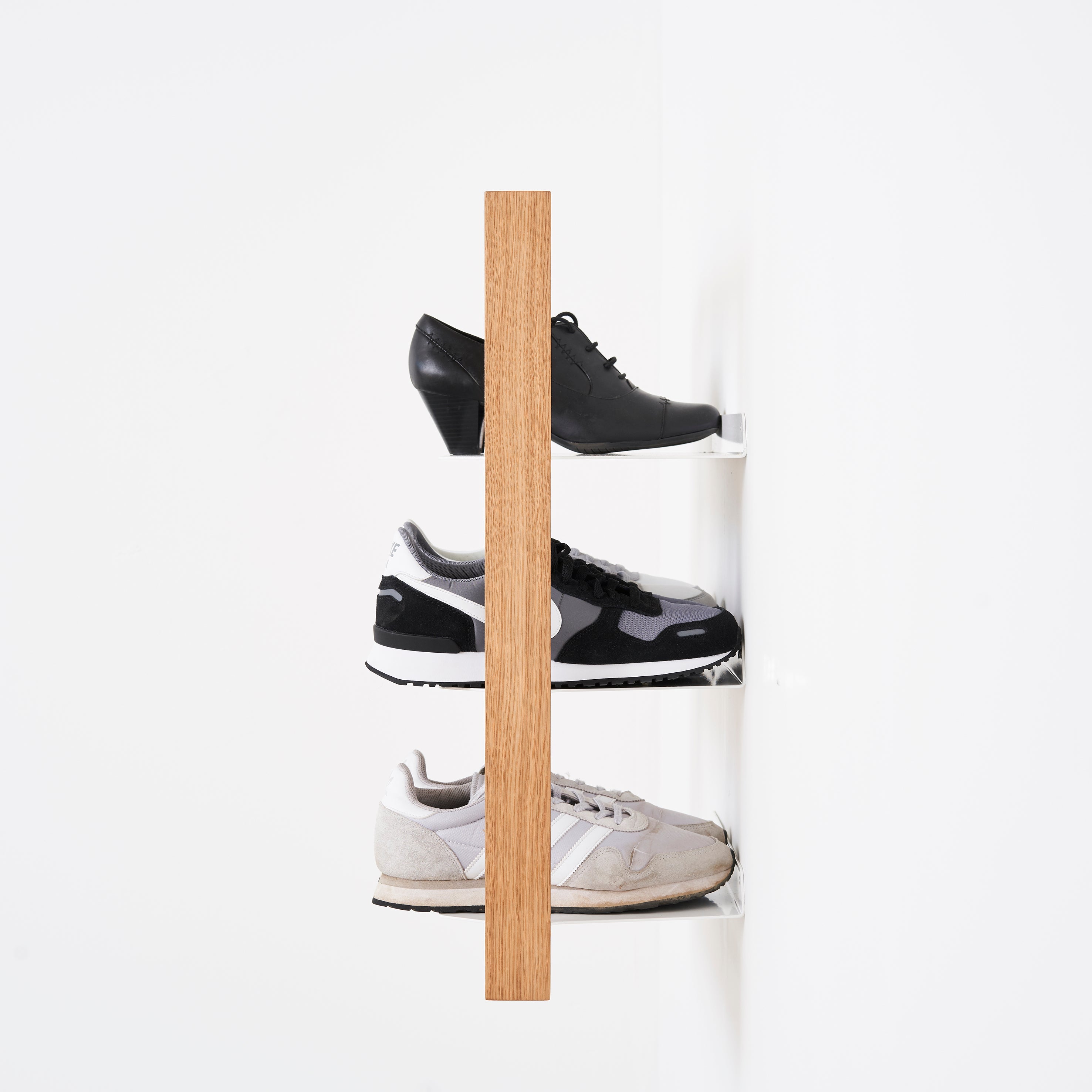 Three pairs of Das kleine bs Basti shoes, featuring black heels, and both black & white and off-white sneakers, are showcased on sleek powder-coated steel shelves with an oak frame against a white wall.