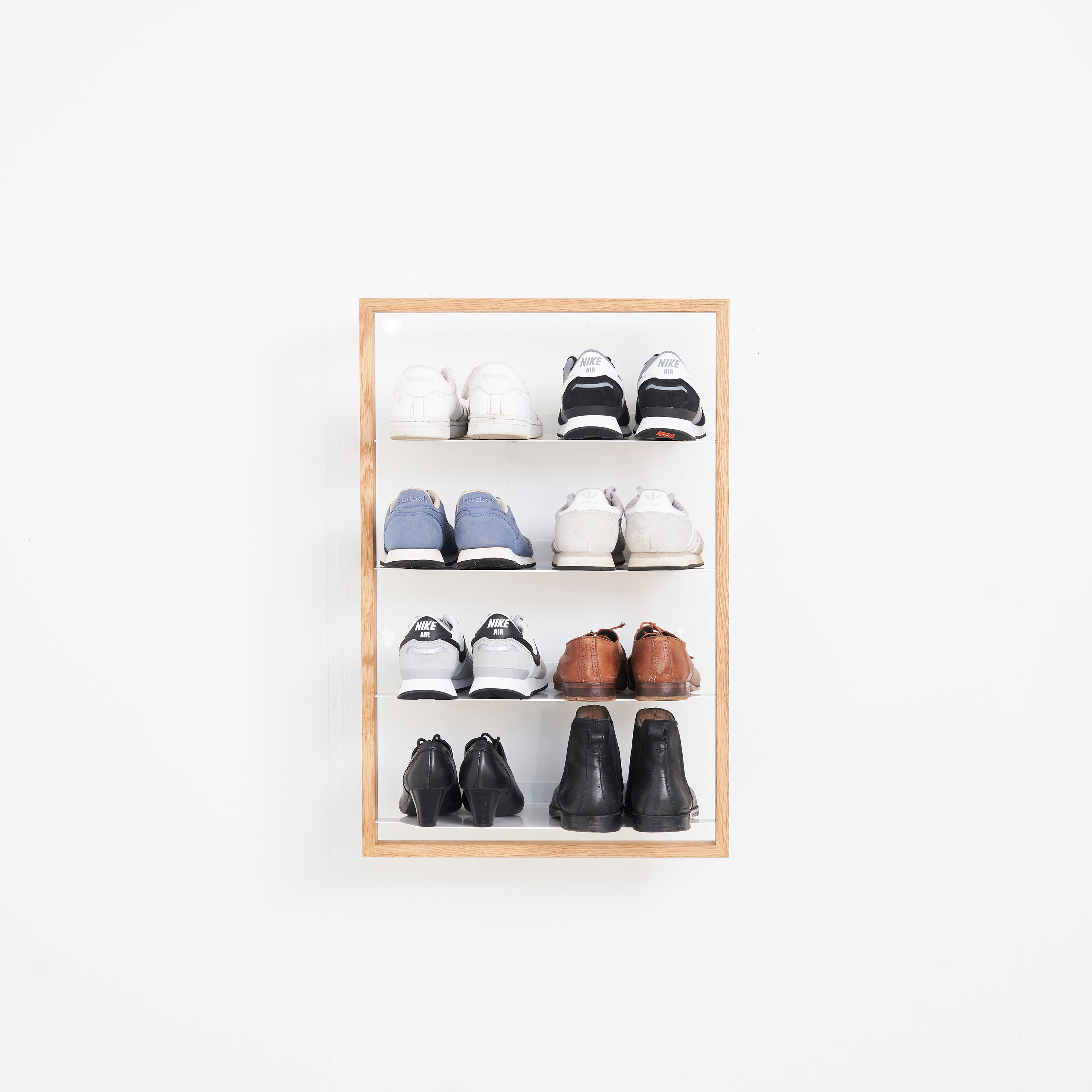 The Basti shoe rack by Das kleine b, with an oak frame and powder-coated steel shelves, is mounted on a white wall. It stylishly displays four pairs of shoes: white sneakers at the top, followed by blue sneakers, brown dress shoes, and black dress shoes at the bottom.