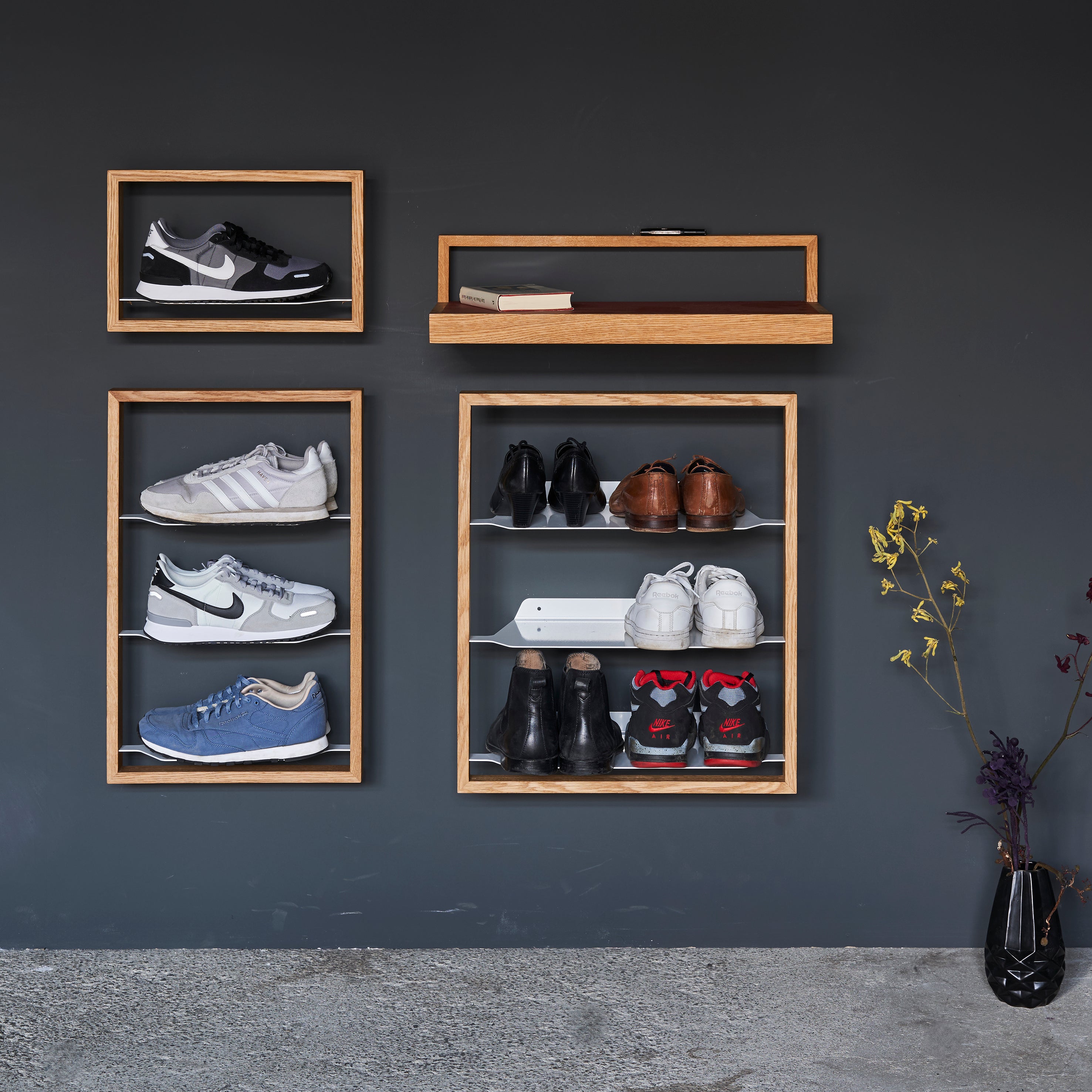 The Basti wall-mounted wooden shoe rack by Das kleine b showcases shoes with its elegant oak frame against a dark wall. A small shelf holds a framed photo, while a floor vase with dried flowers adds decor to this minimalist setup.