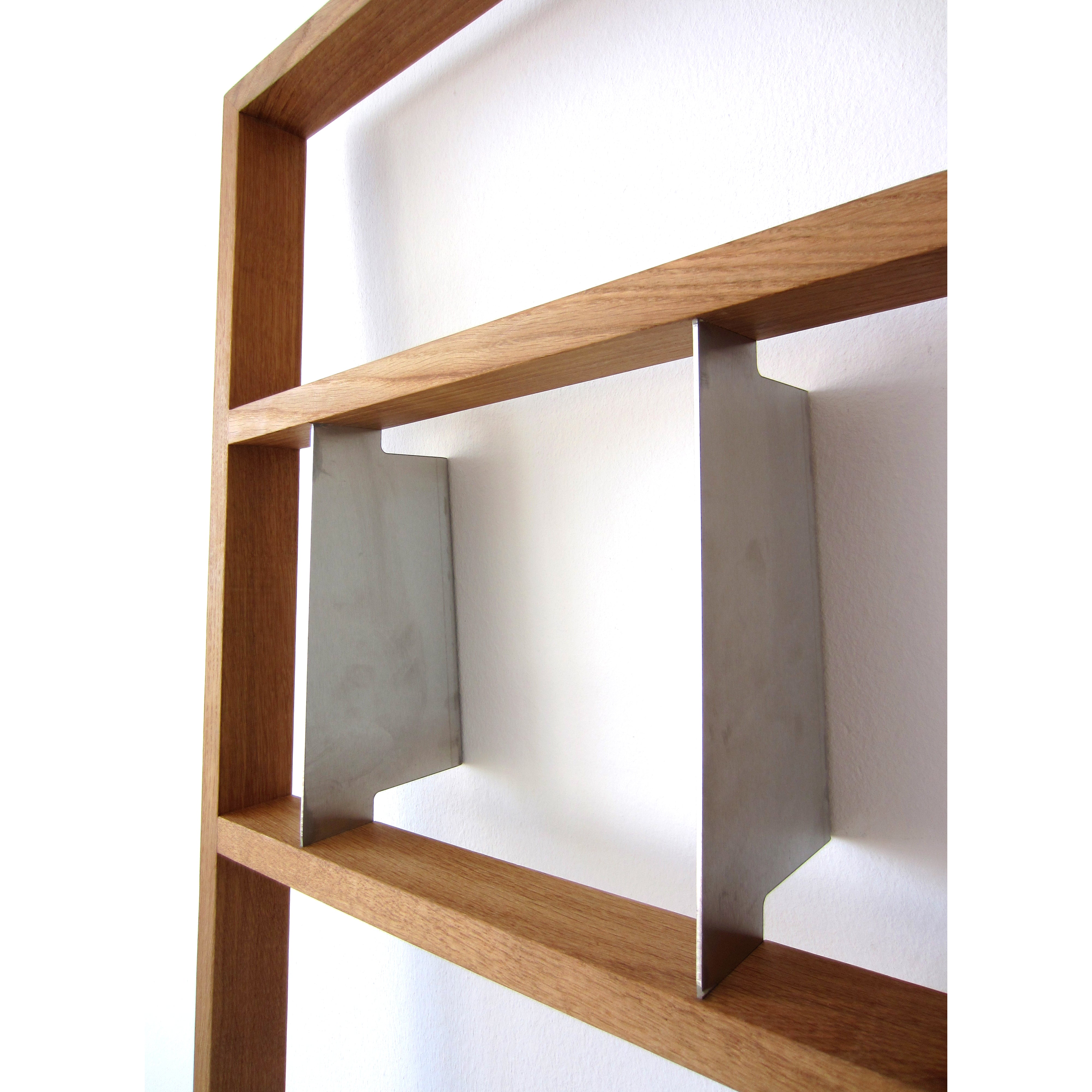 A close-up of the Bertold shelf by Das kleine b highlights its oak and stainless steel design, featuring metal dividers. The empty shelves emphasize the contrast between natural wood grain and sleek metal. Shot from an angle, the image adds depth to the composition.