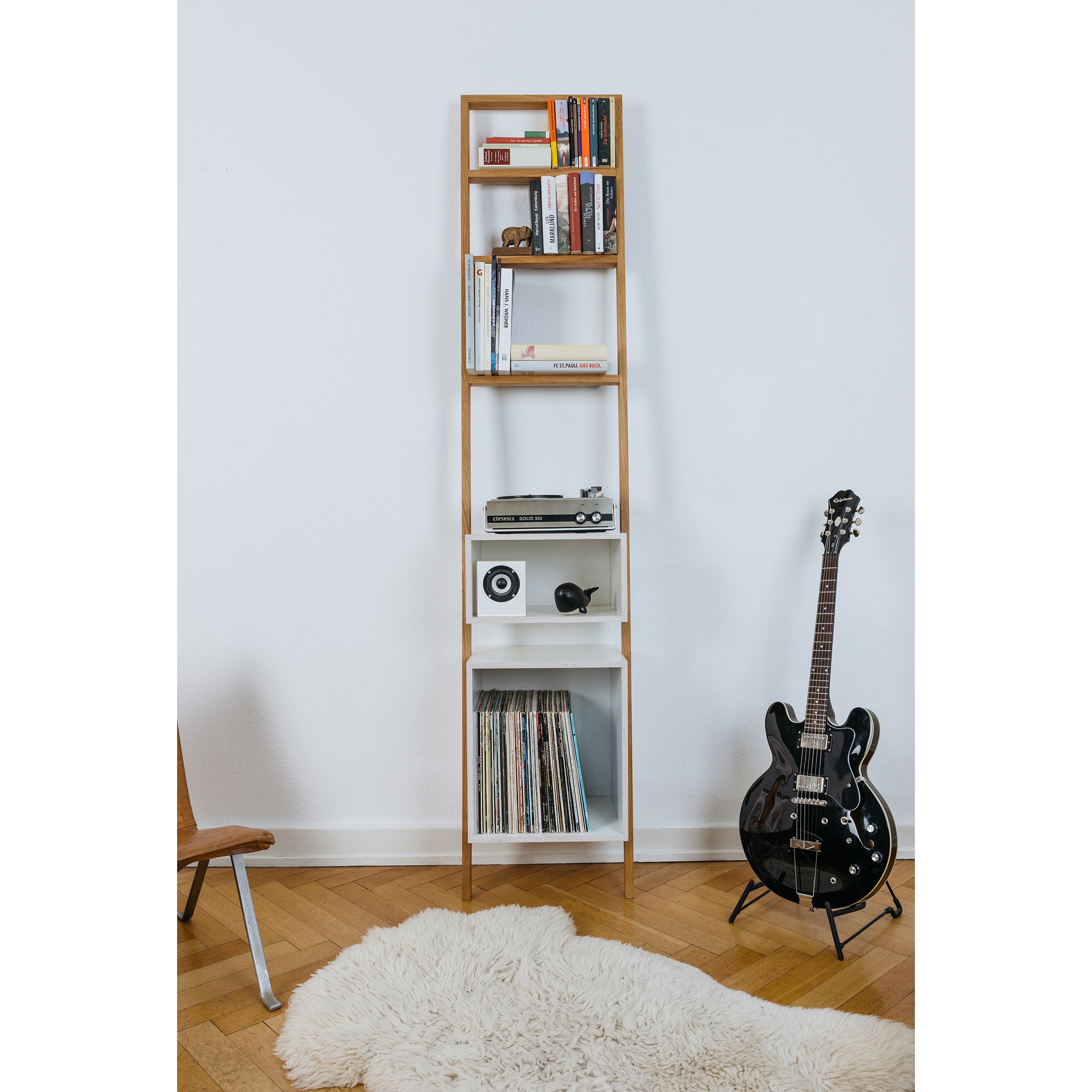 The Brecht ladder-style shelf from Das kleine b, against a white wall, holds books, a camera, a globe, and vinyl records as space-saving furniture. Nearby, an electric guitar stands over a white furry rug on the wooden floor, with the edge of a chair in view.