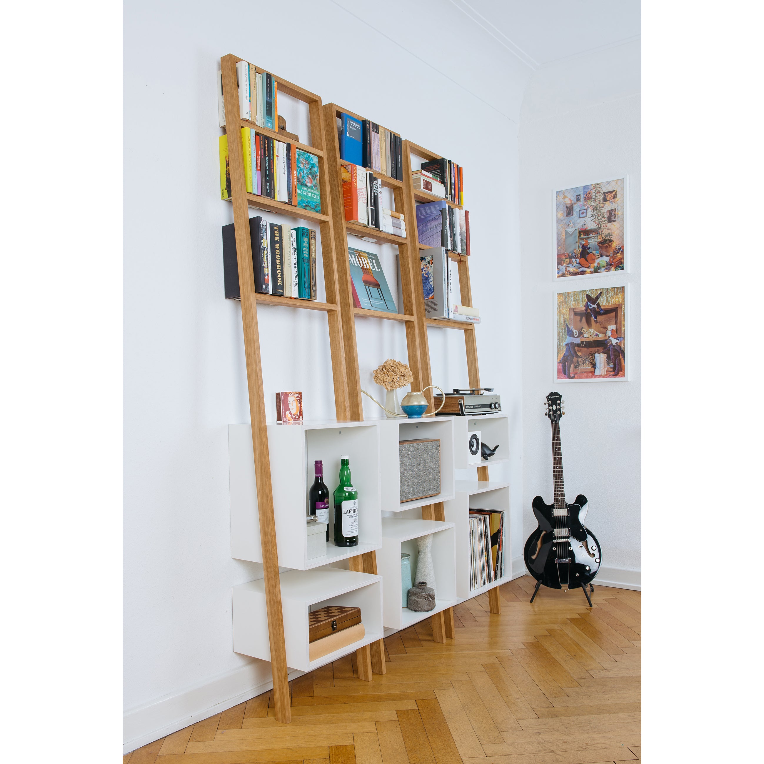 A stylish Brecht ladder rack by Das kleine b showcases books, decor, wine, and a vintage radio against the wall. Nearby rests a black electric guitar on herringbone wood flooring. Modern design paintings complete the look.