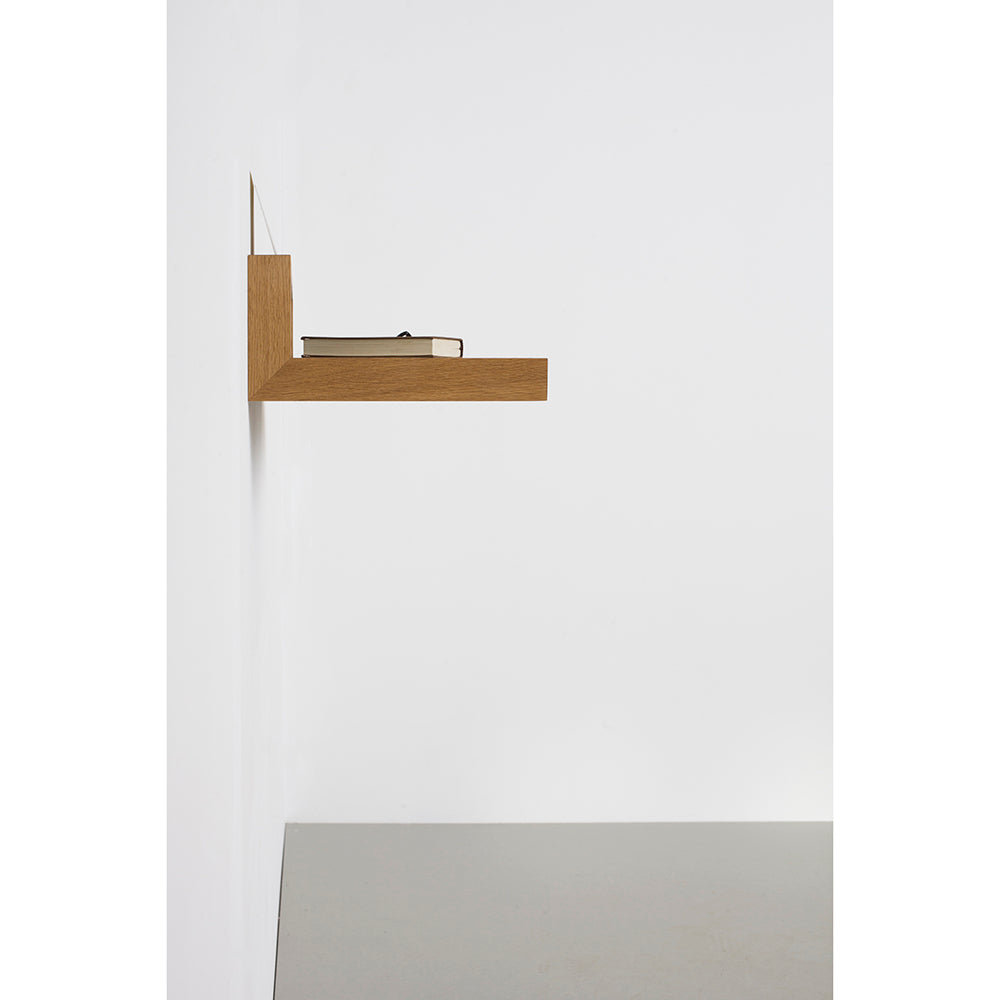 The Buck shelf by Das kleine b, a minimalist wooden storage piece, is mounted on a white wall. A closed beige book rests on it while it hovers gracefully above a light gray linoleum surface.