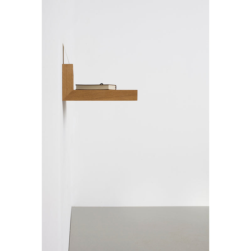 The Buck shelf by Das kleine b, a minimalist wooden storage piece, is mounted on a white wall. A closed beige book rests on it while it hovers gracefully above a light gray linoleum surface.