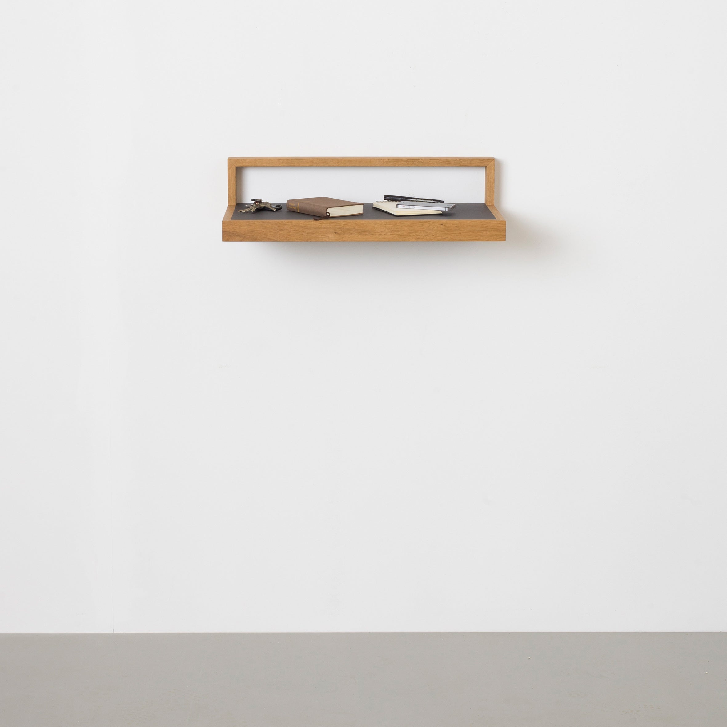 The Buck wooden floating shelf by Das kleine b on a white wall holds a notebook, closed book, and eyeglasses. This versatile piece sits above the gray linoleum surface for a minimalist, uncluttered look.