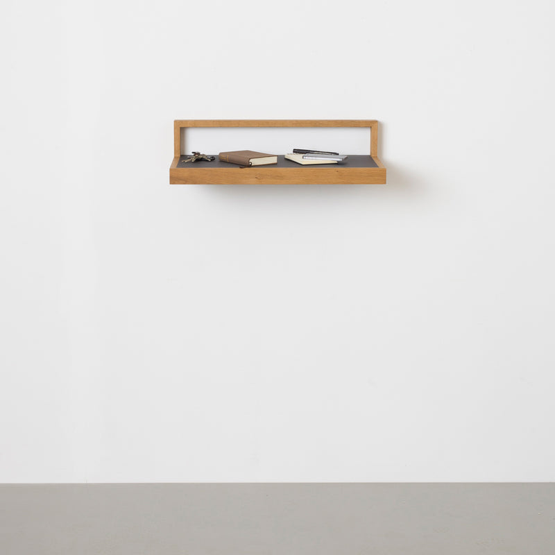 The Buck wooden floating shelf by Das kleine b on a white wall holds a notebook, closed book, and eyeglasses. This versatile piece sits above the gray linoleum surface for a minimalist, uncluttered look.