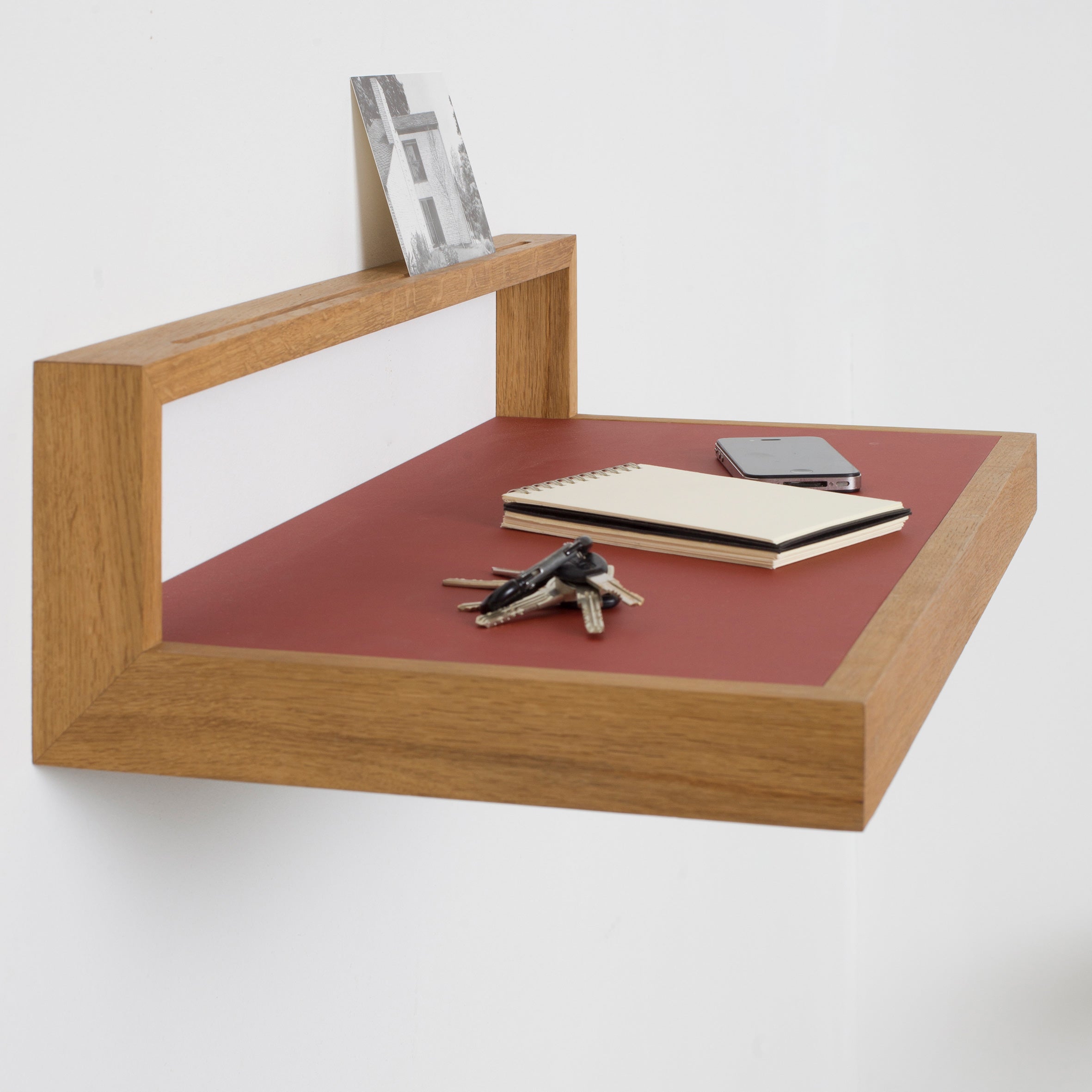 The Buck shelf by Das kleine b, featuring a wooden design with a red linoleum surface, is mounted on a white wall. It holds a photo, notebook, smartphone, and keys.