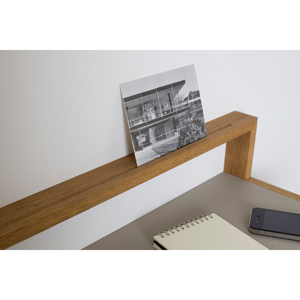 The Buck shelf by Das kleine b features a black and white architectural photo leaning against its wooden frame. Below, a closed notebook and smartphone sit on the linoleum surface. This adaptable piece showcases minimalist and modern design.