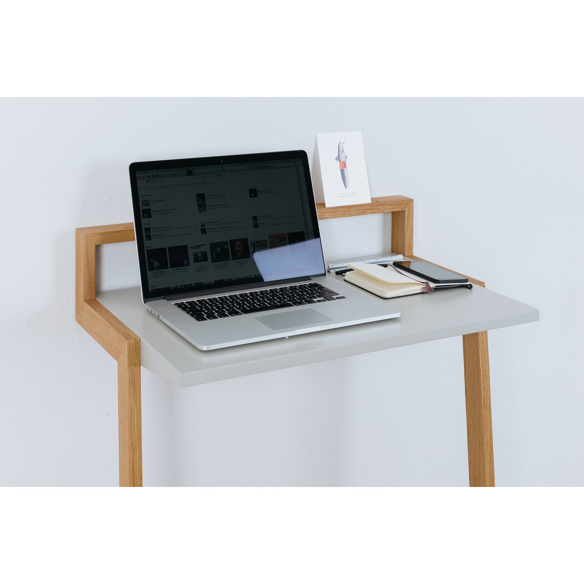 The Buckowski by Das kleine b is a minimalist standing desk with a light gray surface and wooden legs, ideal for a modern mobile workspace. A laptop, closed notebook, pen, smartphone, and illustrated card complete the neat arrangement.