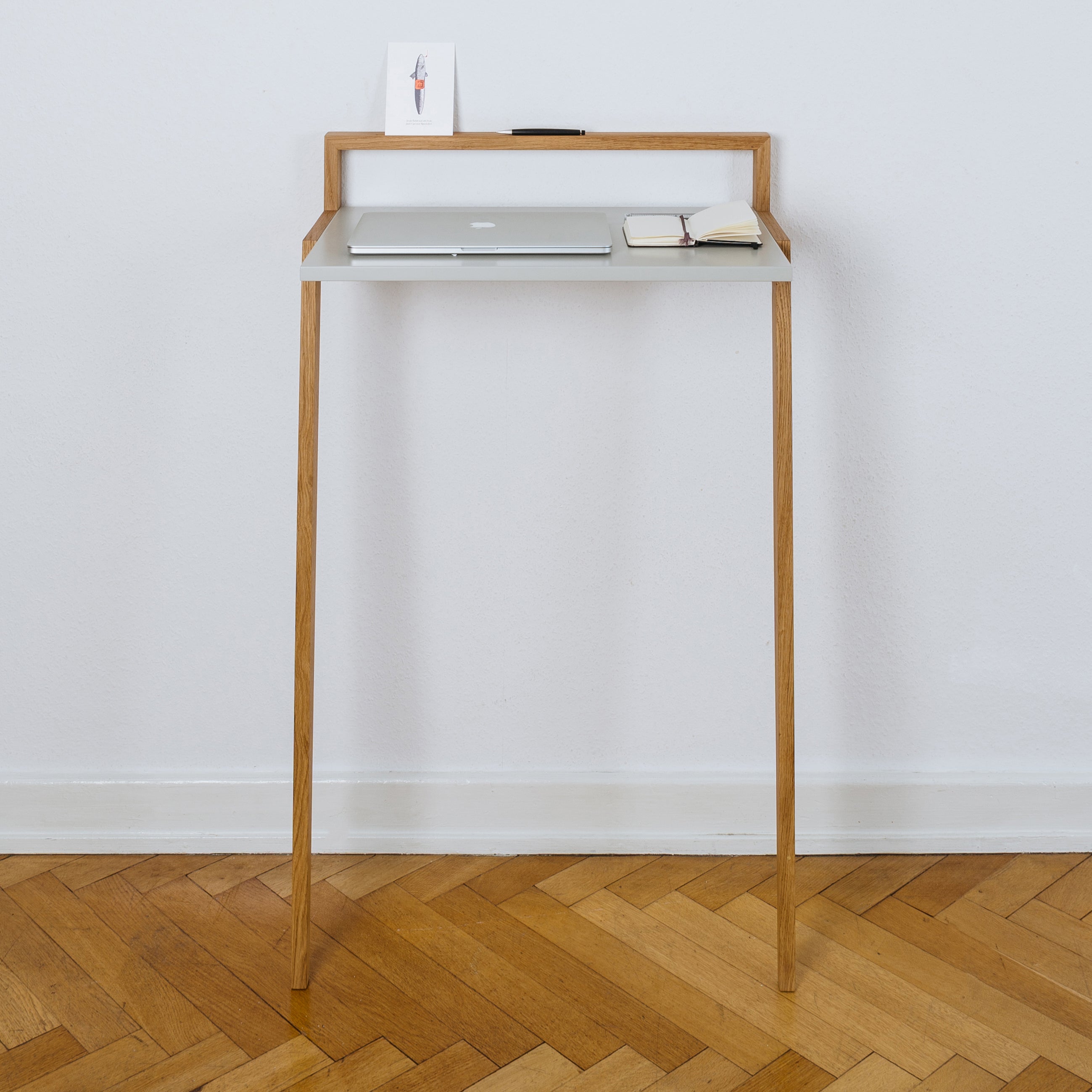 The Buckowski by Das kleine b is a minimalist standing desk with a light wood base and white tabletop, perfect for a modern mobile workspace. It includes a closed laptop, small notebook, and illustrated card and features sleek angled legs on a herringbone-patterned floor.