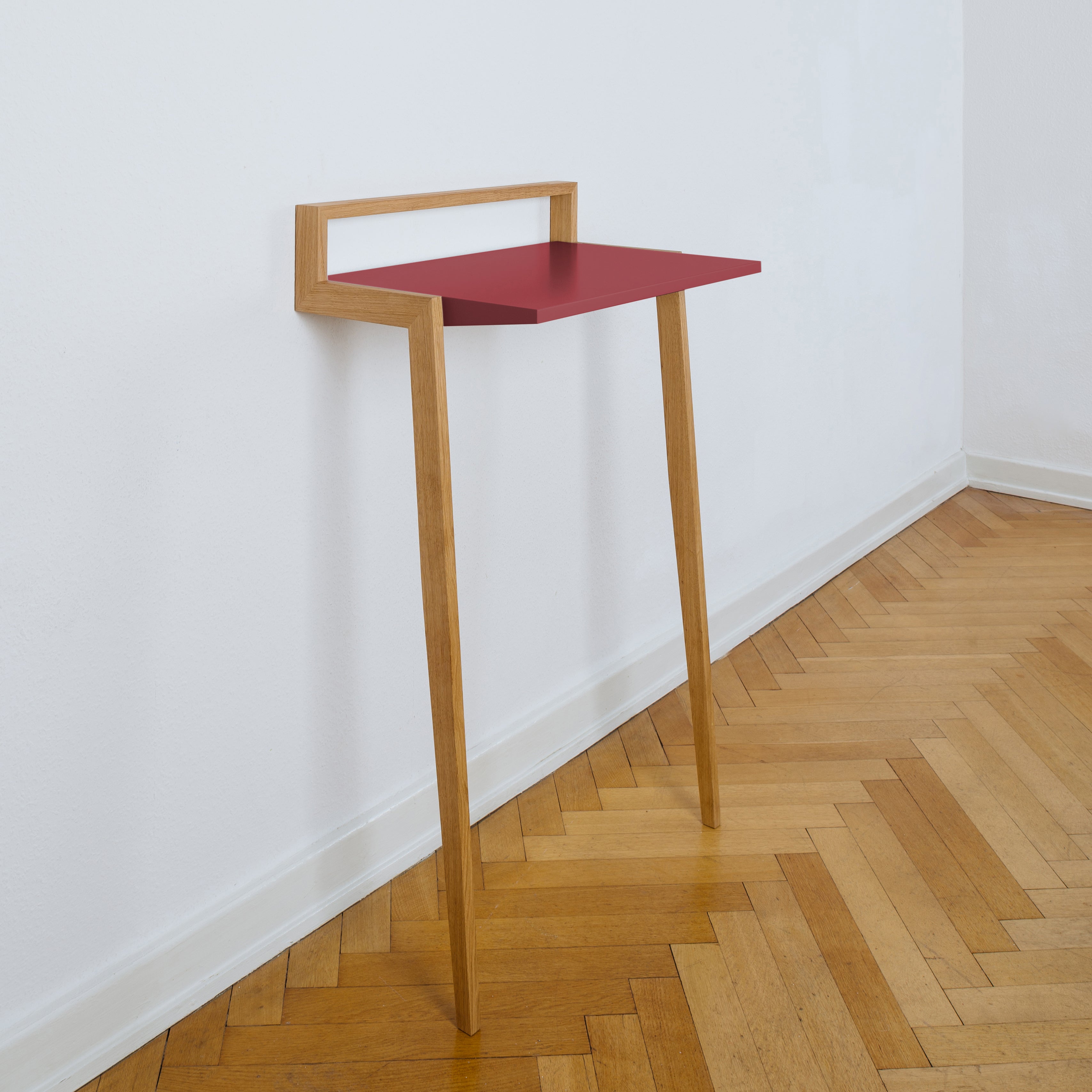 The Buckowski by Das kleine b is a minimalist wooden side table with a red top, attached to a white wall, highlighting its sleek design. It stands on two angled wooden legs and elegantly rests on a herringbone-patterned wooden floor.