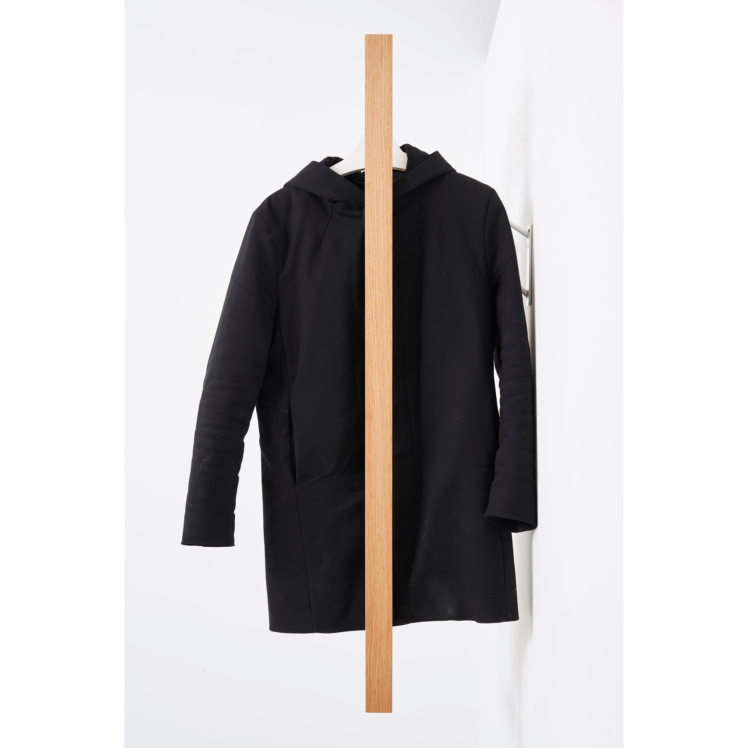 The black Gardero-b coat by Das kleine b hangs on a vertical wooden beam against a white wall, creating symmetrical organization. The beam divides the coat in half, showing only one sleeve side and enhancing its minimalist aesthetic.