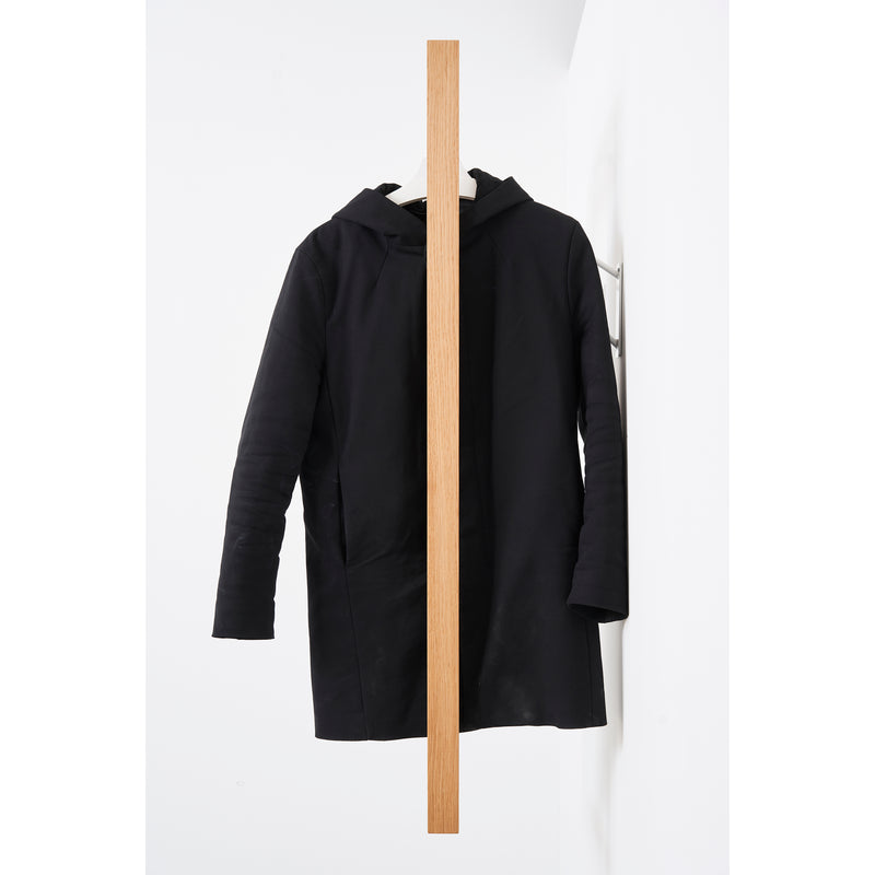 The black Gardero-b coat by Das kleine b hangs on a vertical wooden beam against a white wall, creating symmetrical organization. The beam divides the coat in half, showing only one sleeve side and enhancing its minimalist aesthetic.