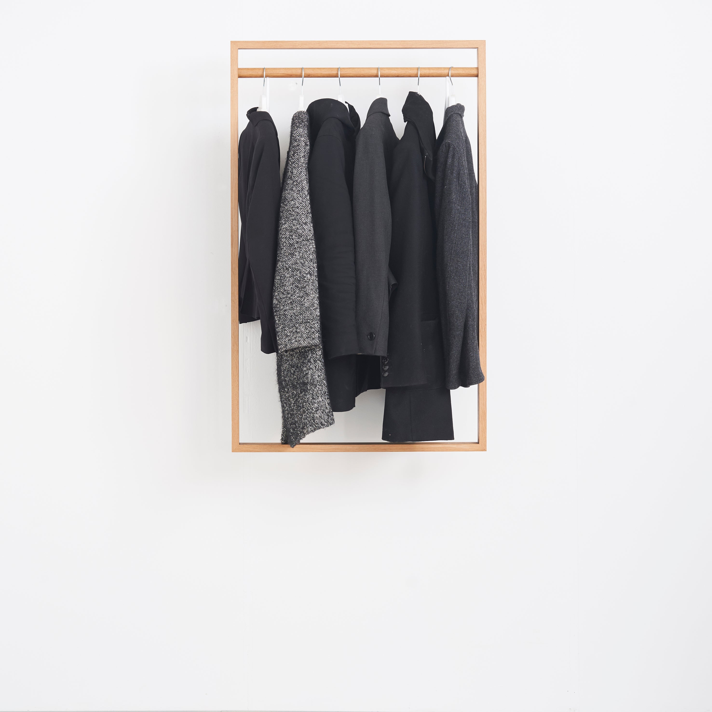 The Gardero-b by Das kleine b displays six dark-colored coats and jackets hung neatly against a white wall. The black and grey garments complement the hall stands simple design, showcasing diverse textures and materials while optimizing wardrobe space.