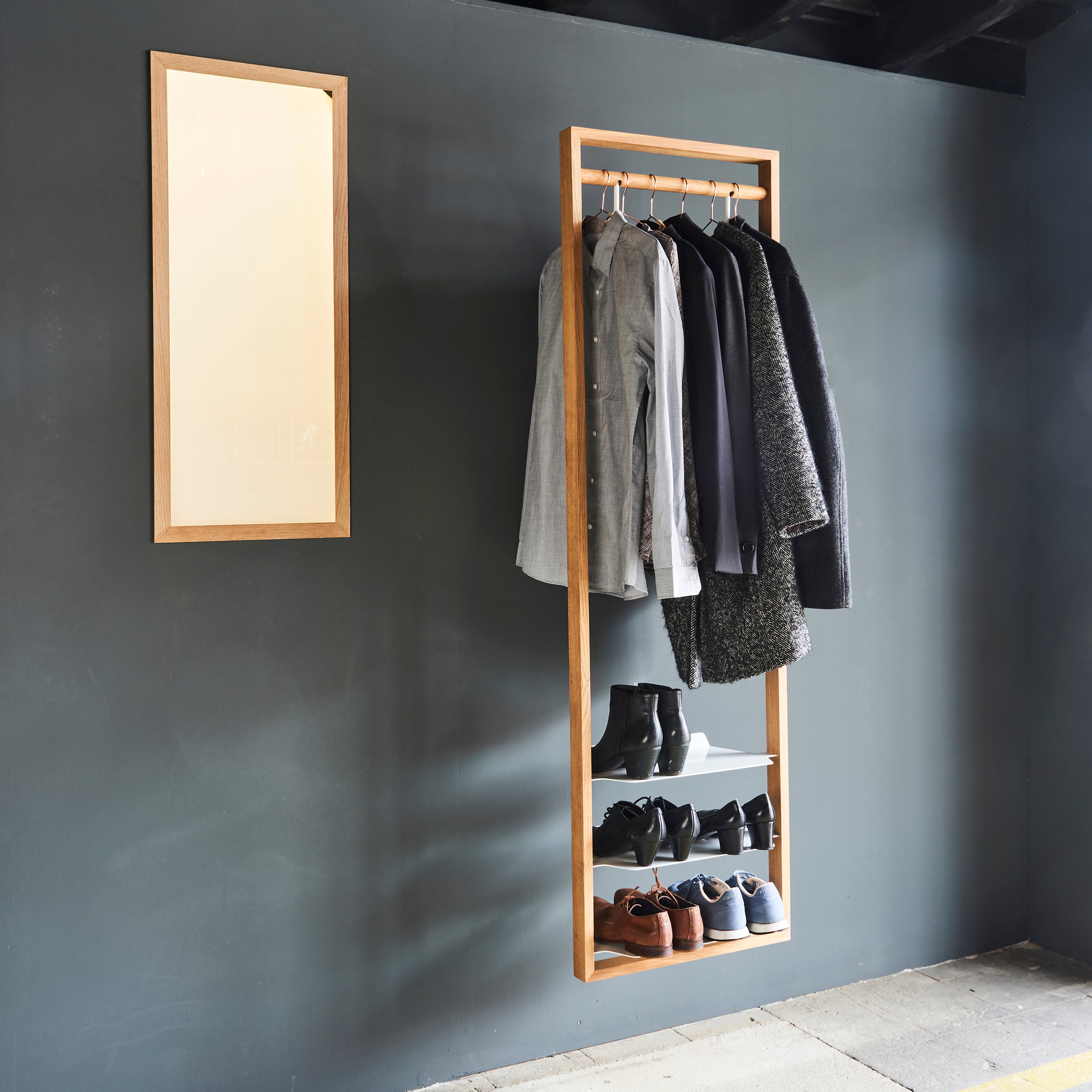 The Gardero-b by Das kleine b elegantly displays shirts, jackets, and diverse footwear on a minimalist wooden hall stand against a dark gray wall. A rectangular mirror with a wooden frame nearby enhances the stylish wardrobe-like setup.