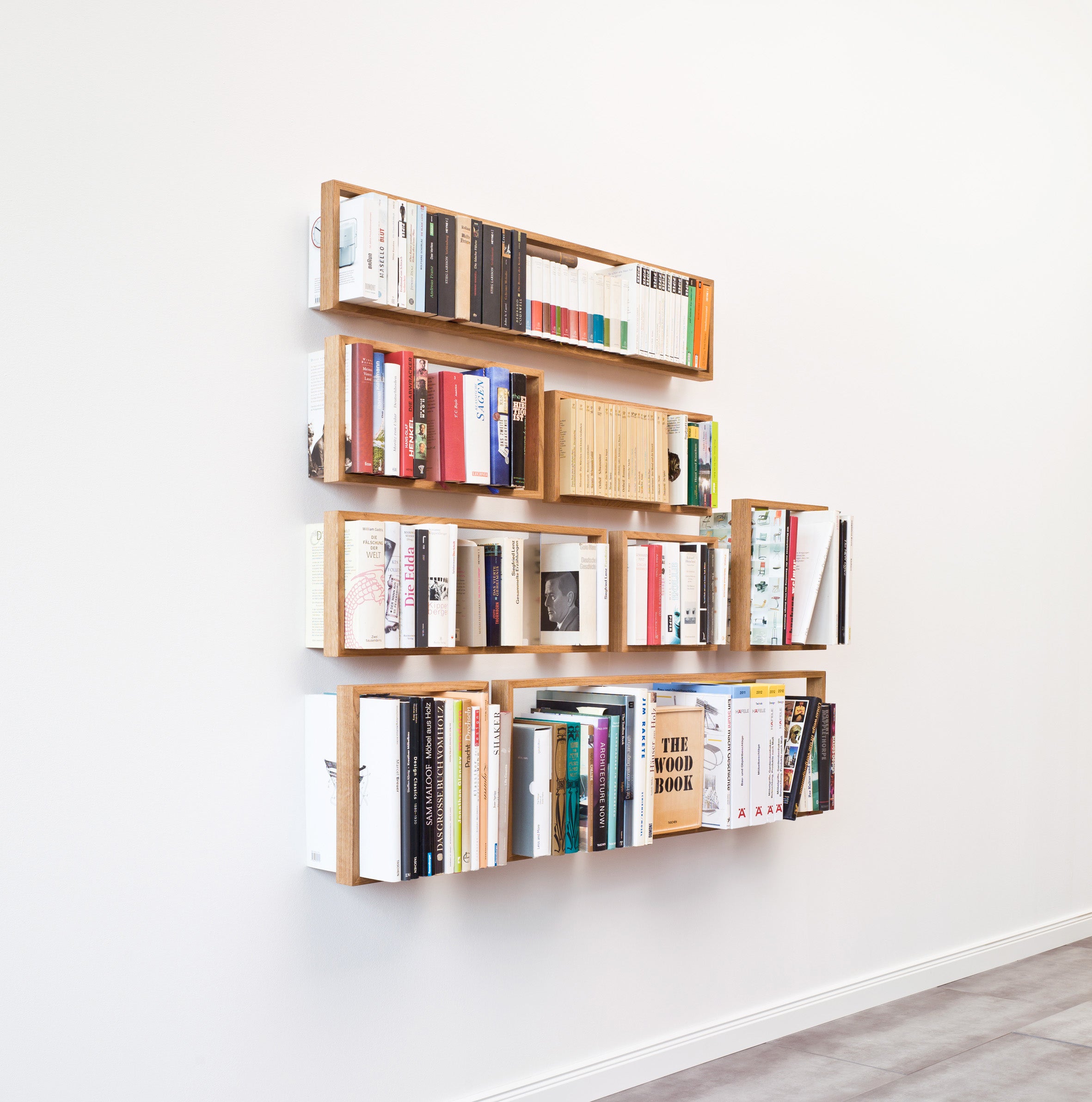 The Das kleine bs 12.2 wooden floating bookshelves elegantly hold books arranged both vertically and horizontally. Set against a plain white wall above a tiled floor, they create a minimalist and orderly effect through the interplay of light and shadows.