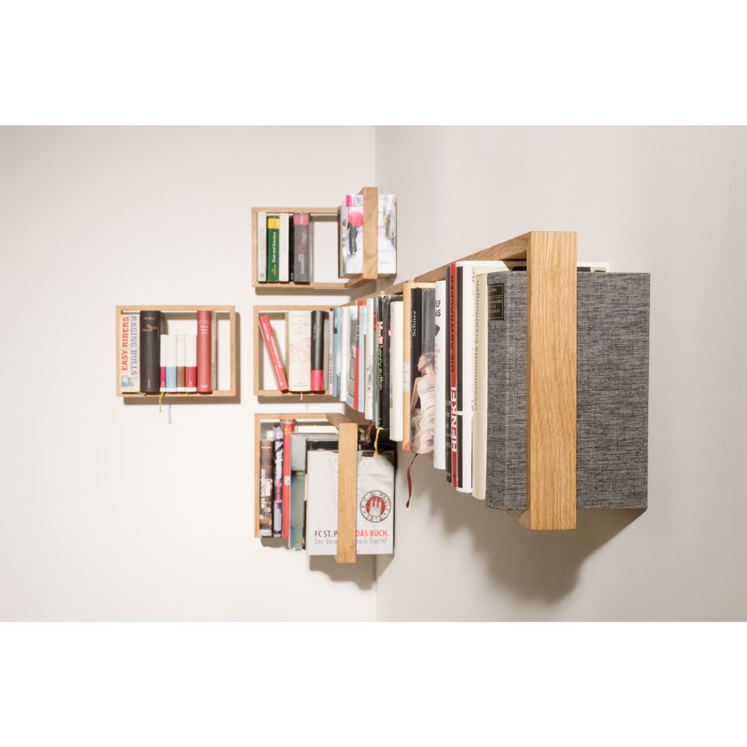 The b 12.2 floating wooden wall-mounted bookshelves by Das kleine b present an artistic display with books arranged in diverse orientations. Set at varying lengths and heights on a white wall, they add a dynamic touch to any room.