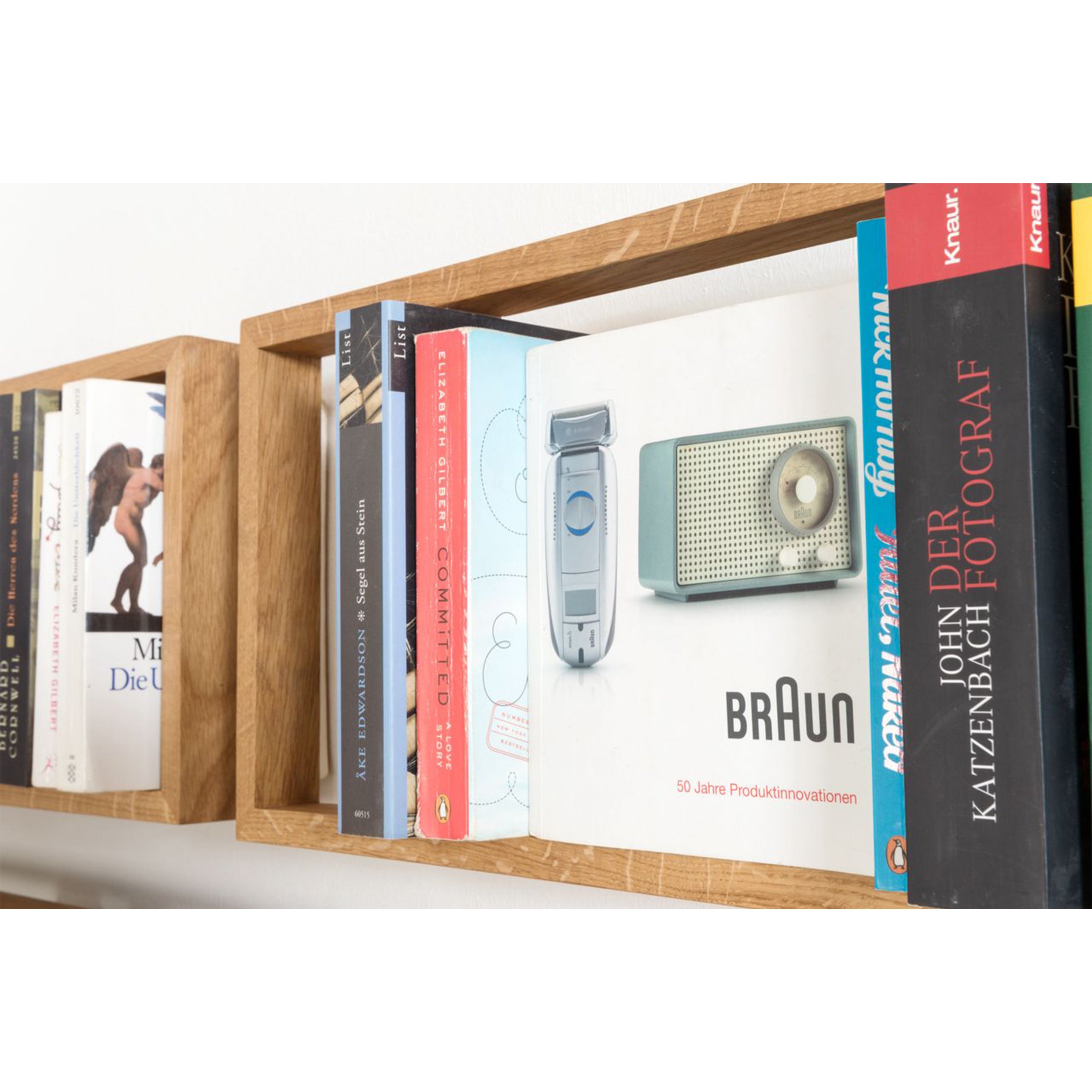 A wooden bookshelf with stainless steel angles showcases colorful books alongside a vintage Braun electric razor box. This vibrant book collection contrasts sharply with the minimalist packaging of the b 8.7 by Das kleine b, blending modern and retro aesthetics.